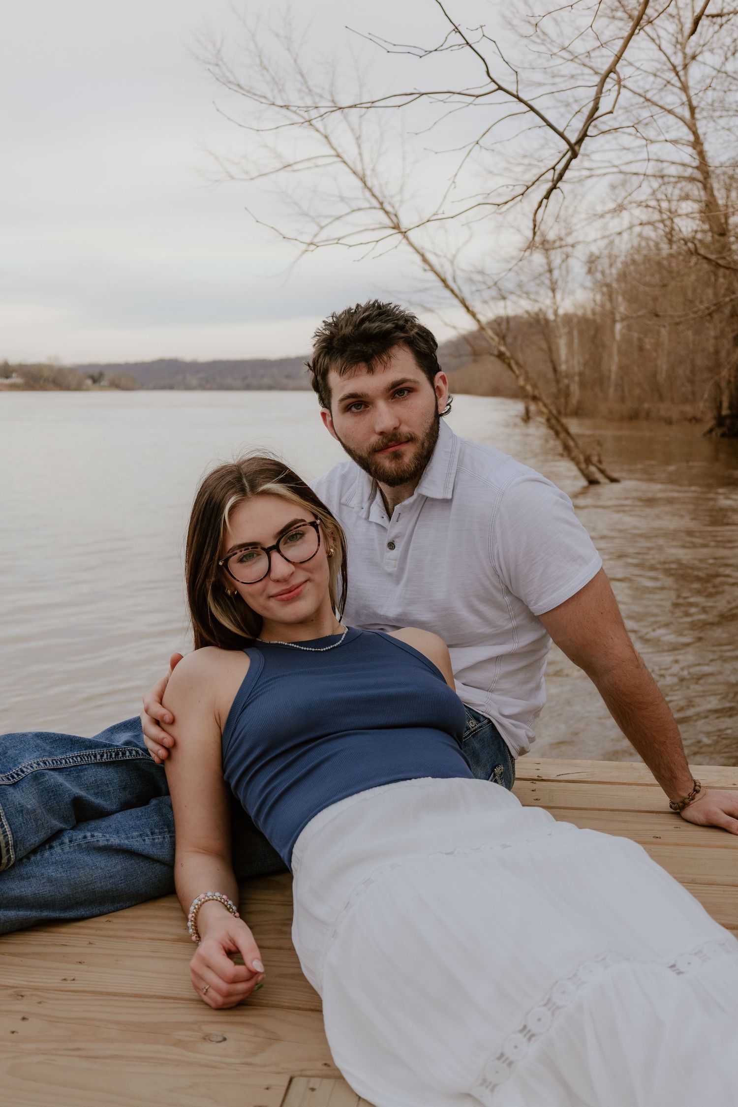 Couple relaxes together on wooden dock by lake during sunset photoshoot in casual attire.