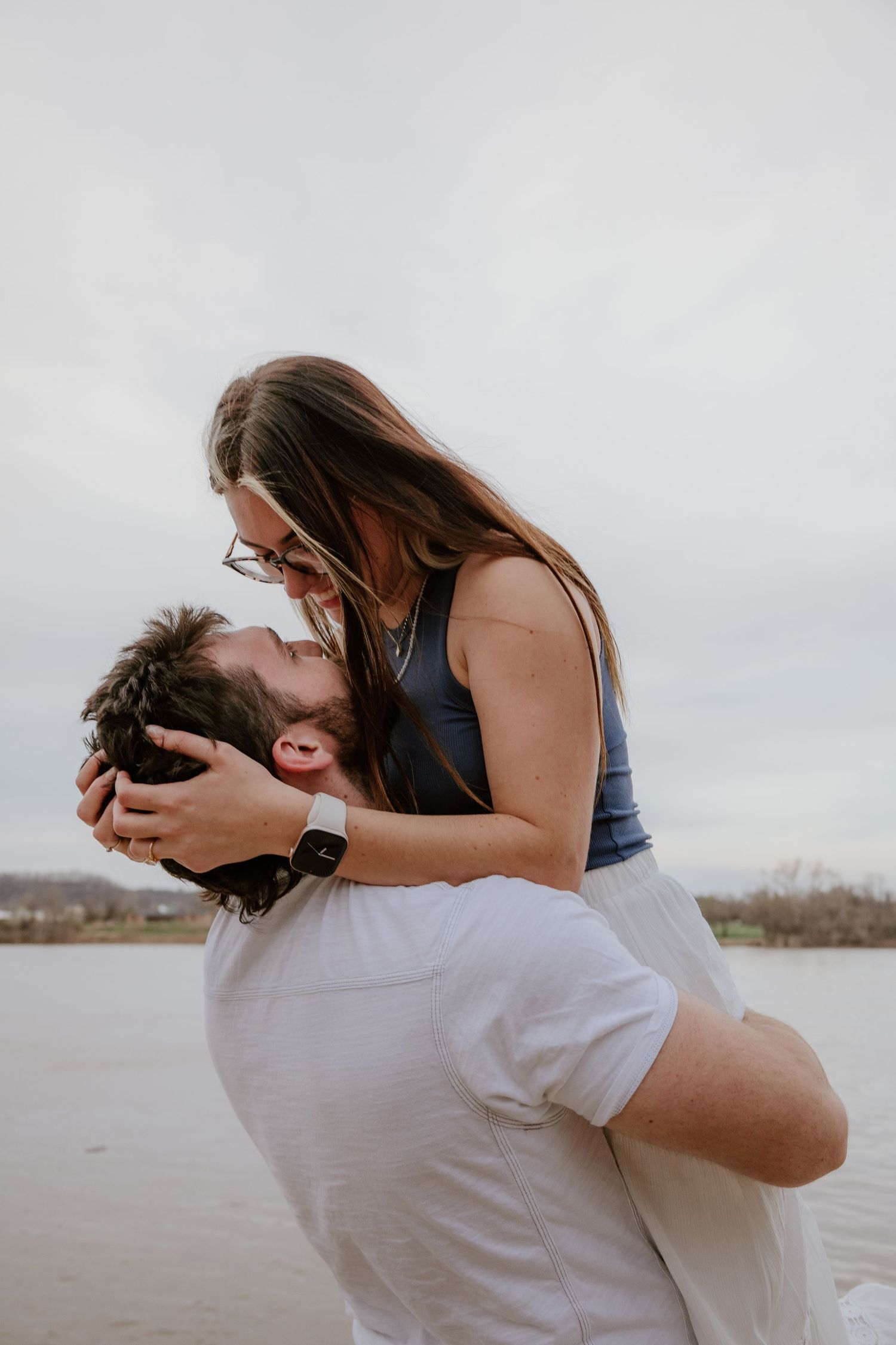 A couple embraces playfully by a lake on an overcast day, creating a romantic series of candid moments.