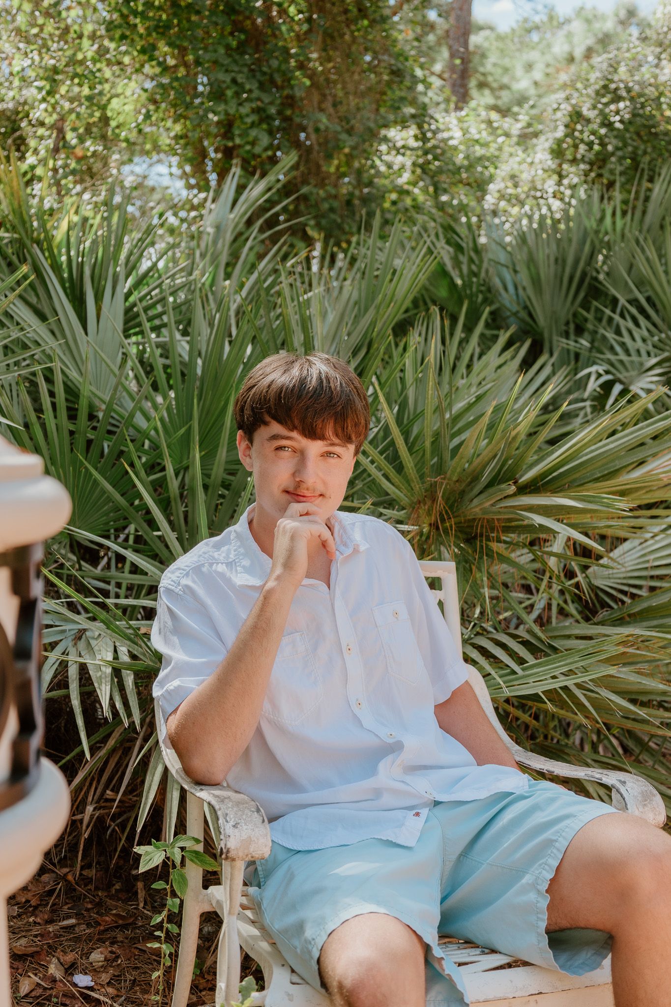 A person in a white shirt and light blue shorts sits casually near tropical palm plants in an outdoor setting.