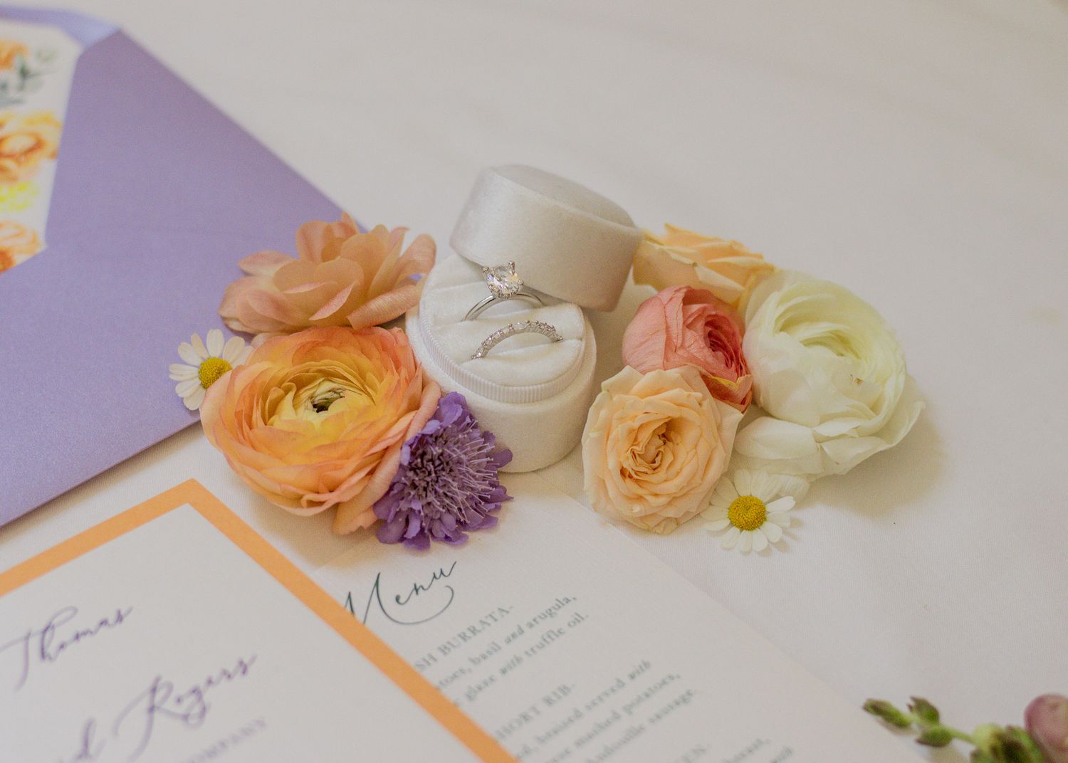 Wedding invitation with peach and cream colored ranunculus flowers and rings on a purple background.
