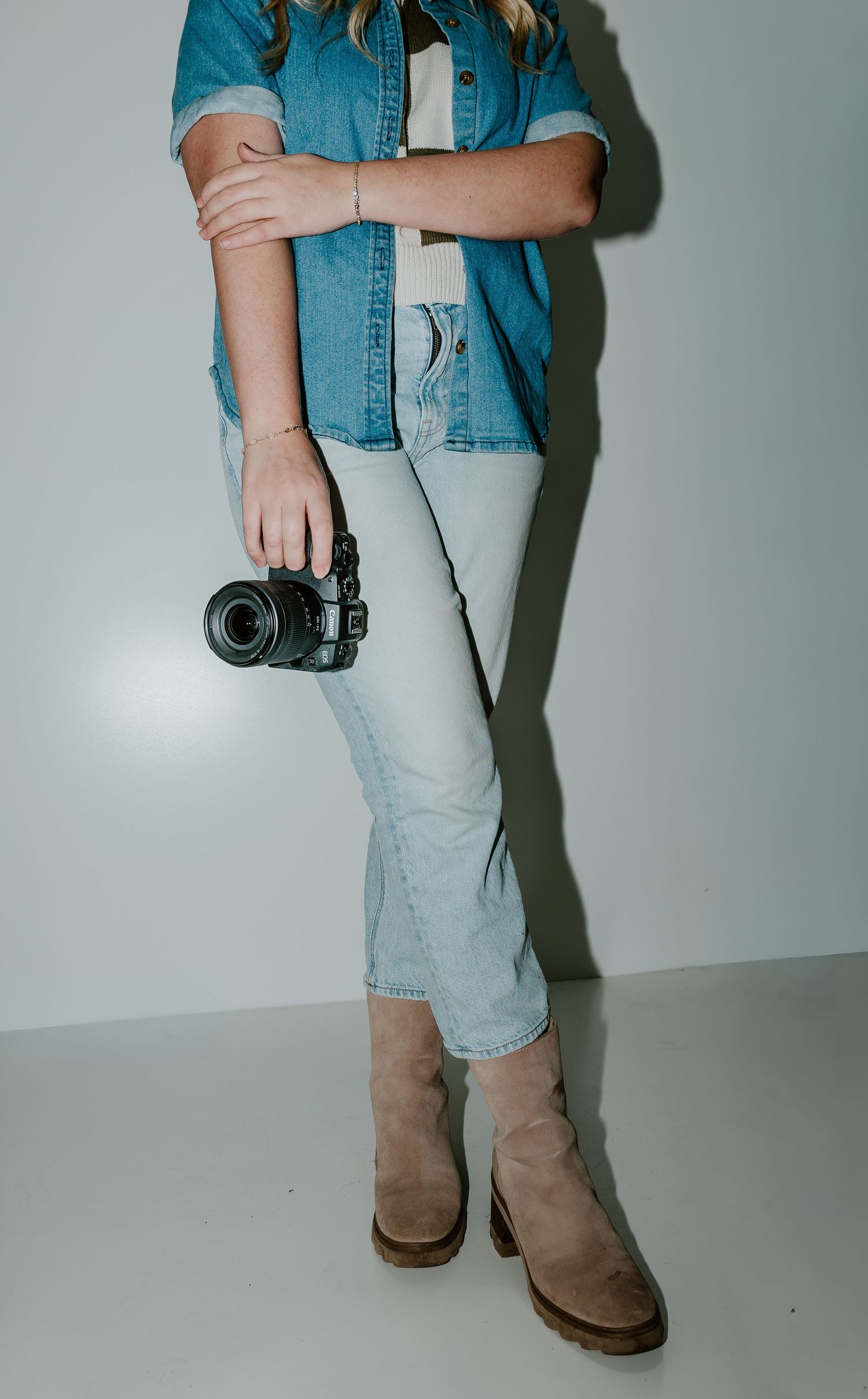 A person wearing light wash jeans and a denim shirt holds a vintage camera against a plain gray background.