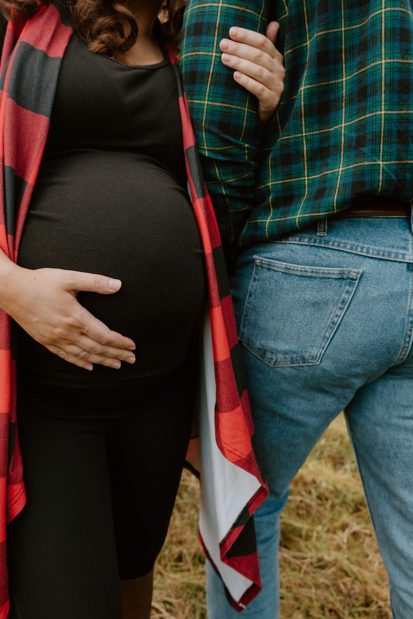 Couple in plaid and denim holding hands during pregnancy photoshoot outdoors.
