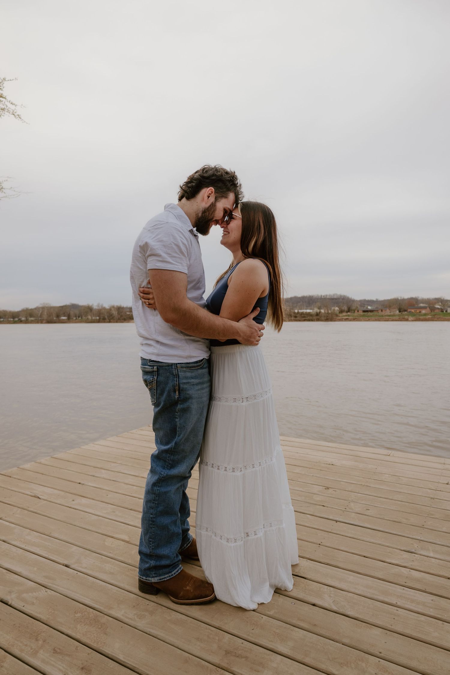 A couple shares an intimate moment on a wooden dock overlooking a serene lake on an overcast day.