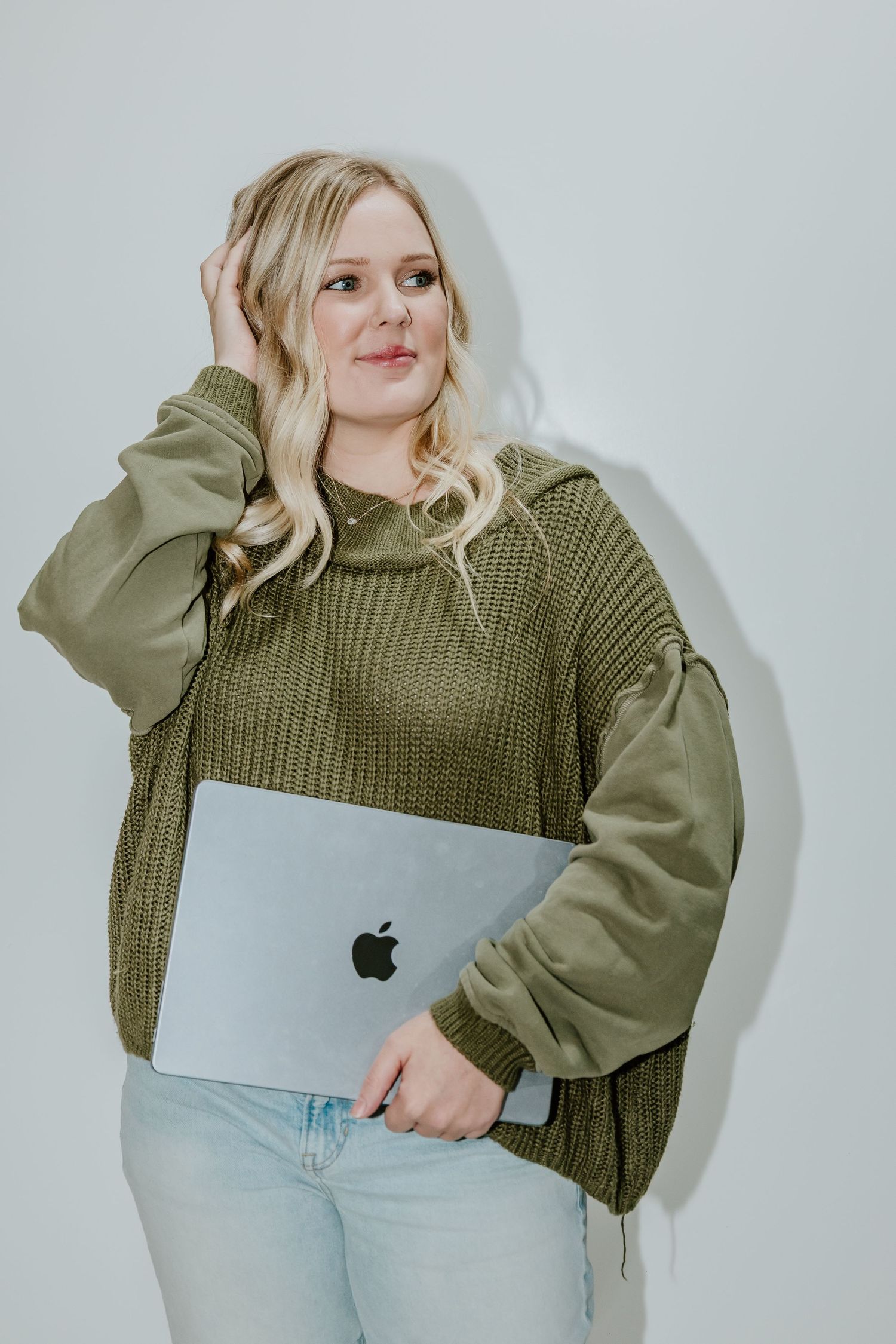 A person in an olive green sweater and light jeans holds a MacBook laptop against a white background.