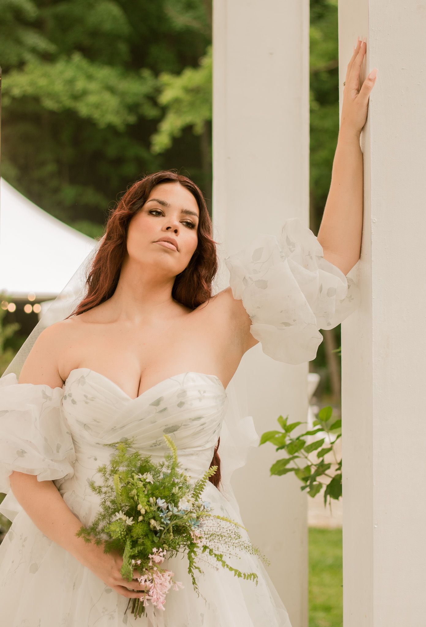 A bride in a white off-shoulder wedding dress poses gracefully against white pillars in an outdoor garden setting.