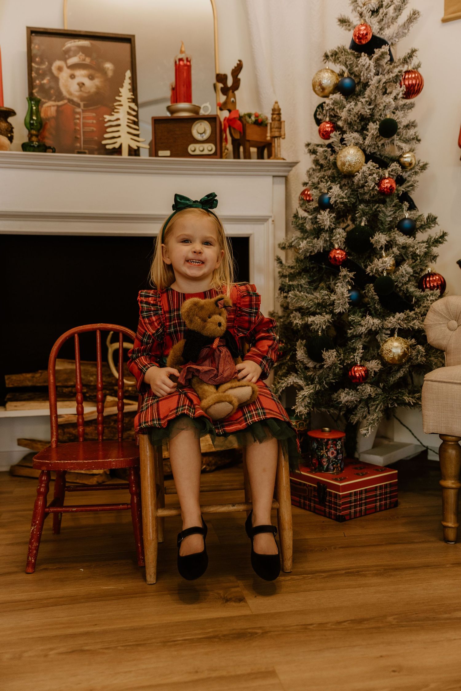 A child in a plaid holiday dress sits on red wooden sleds arranged in a row next to a decorated Christmas tree.