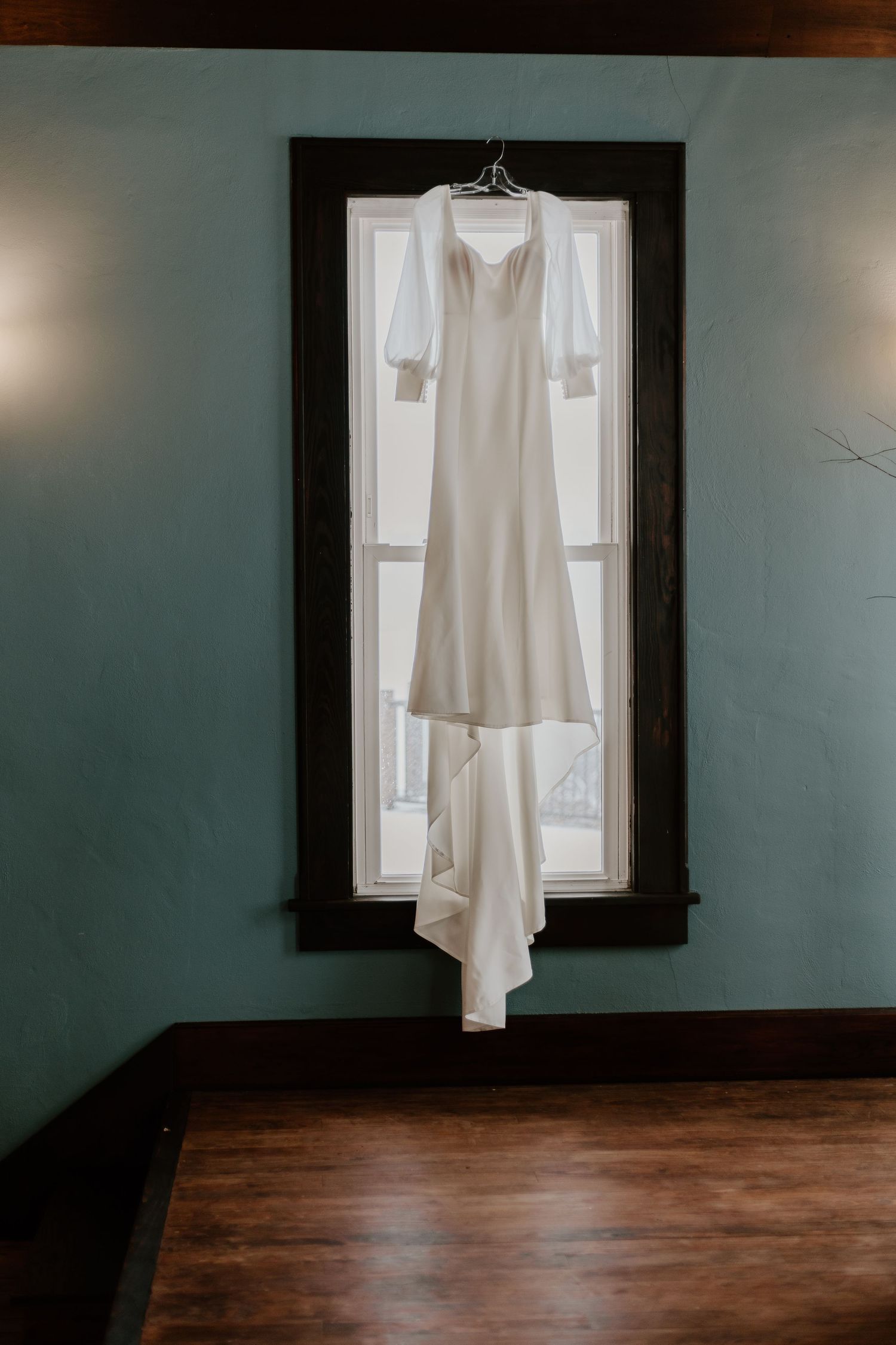 Elegant white wedding dress hanging in front of window with dark wood trim and teal walls.
