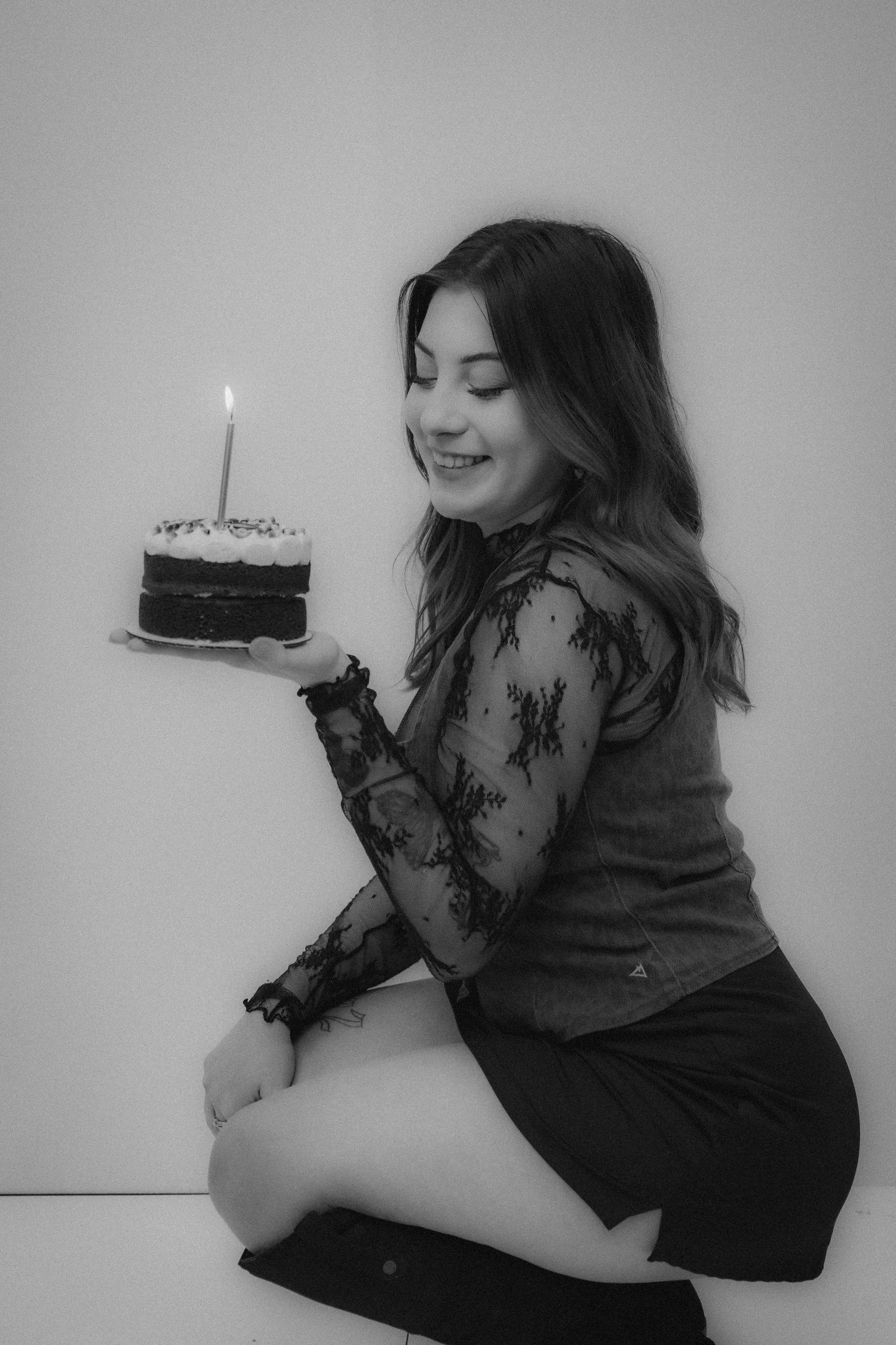 A woman in black lace sleeves lies on a white surface holding a slice of chocolate cake with a candle.