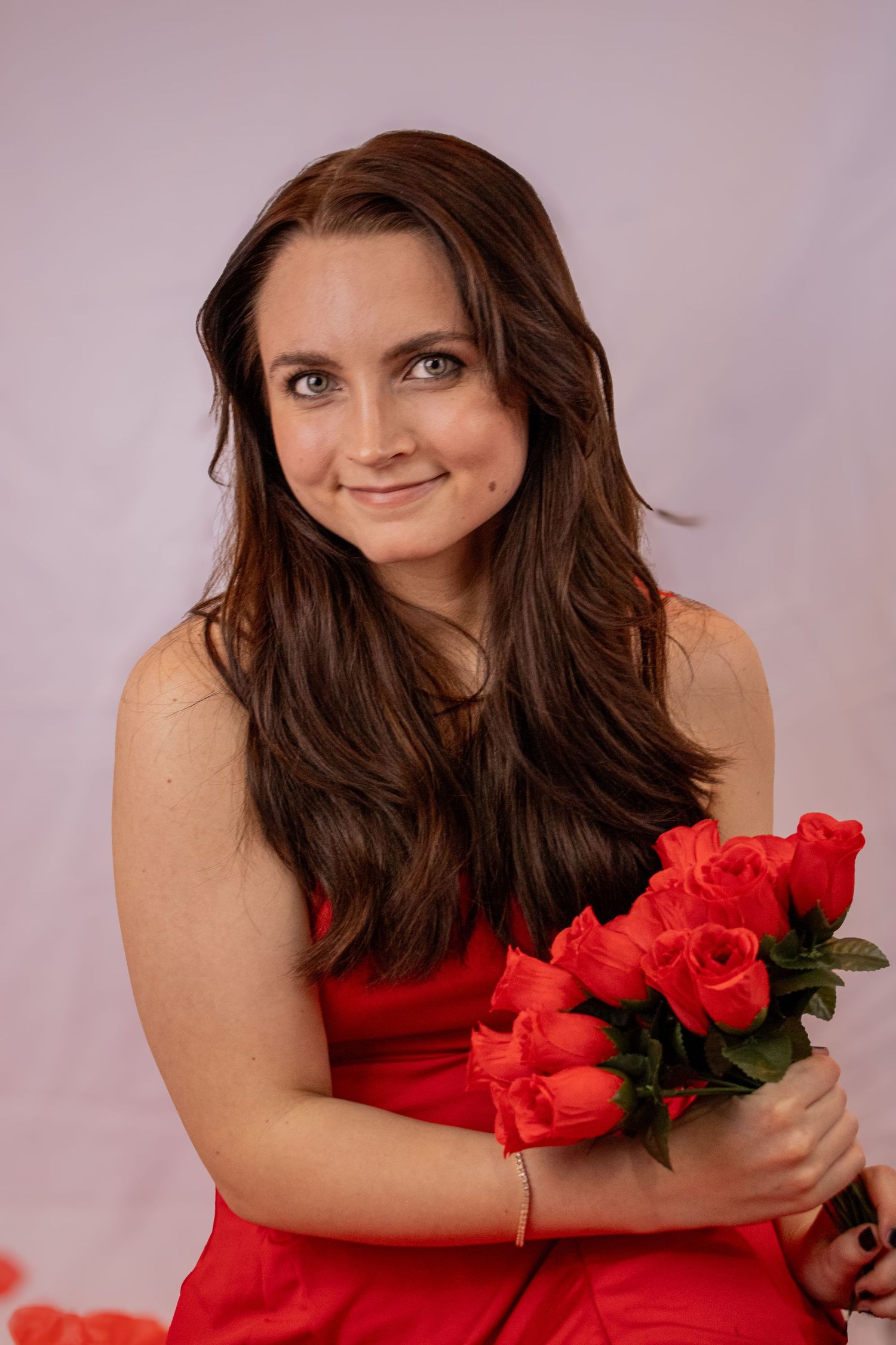 A woman in a red dress poses with vibrant red roses against a soft pink studio backdrop.