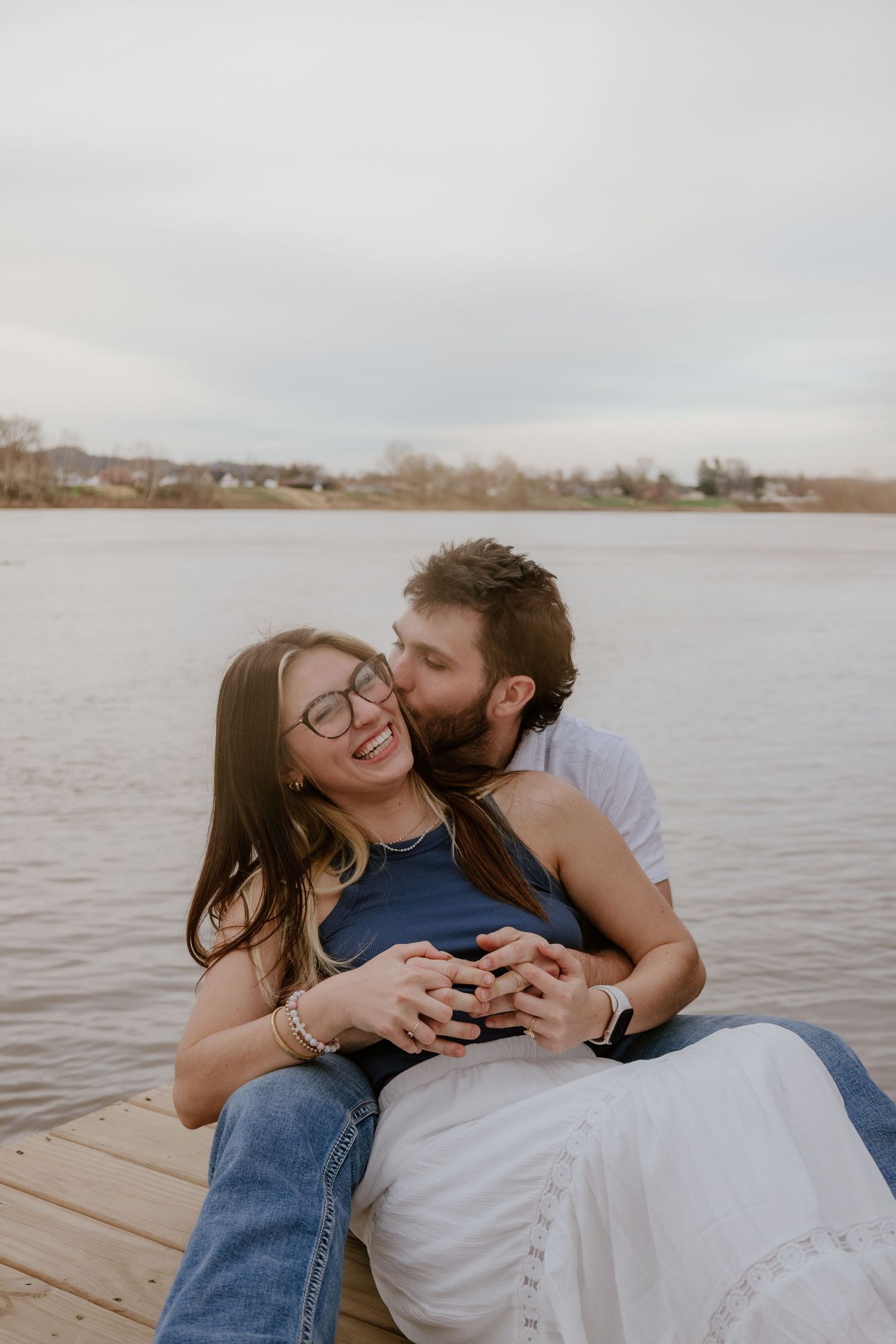 A couple shares intimate moments while relaxing on a wooden dock by a serene lake on an overcast day.