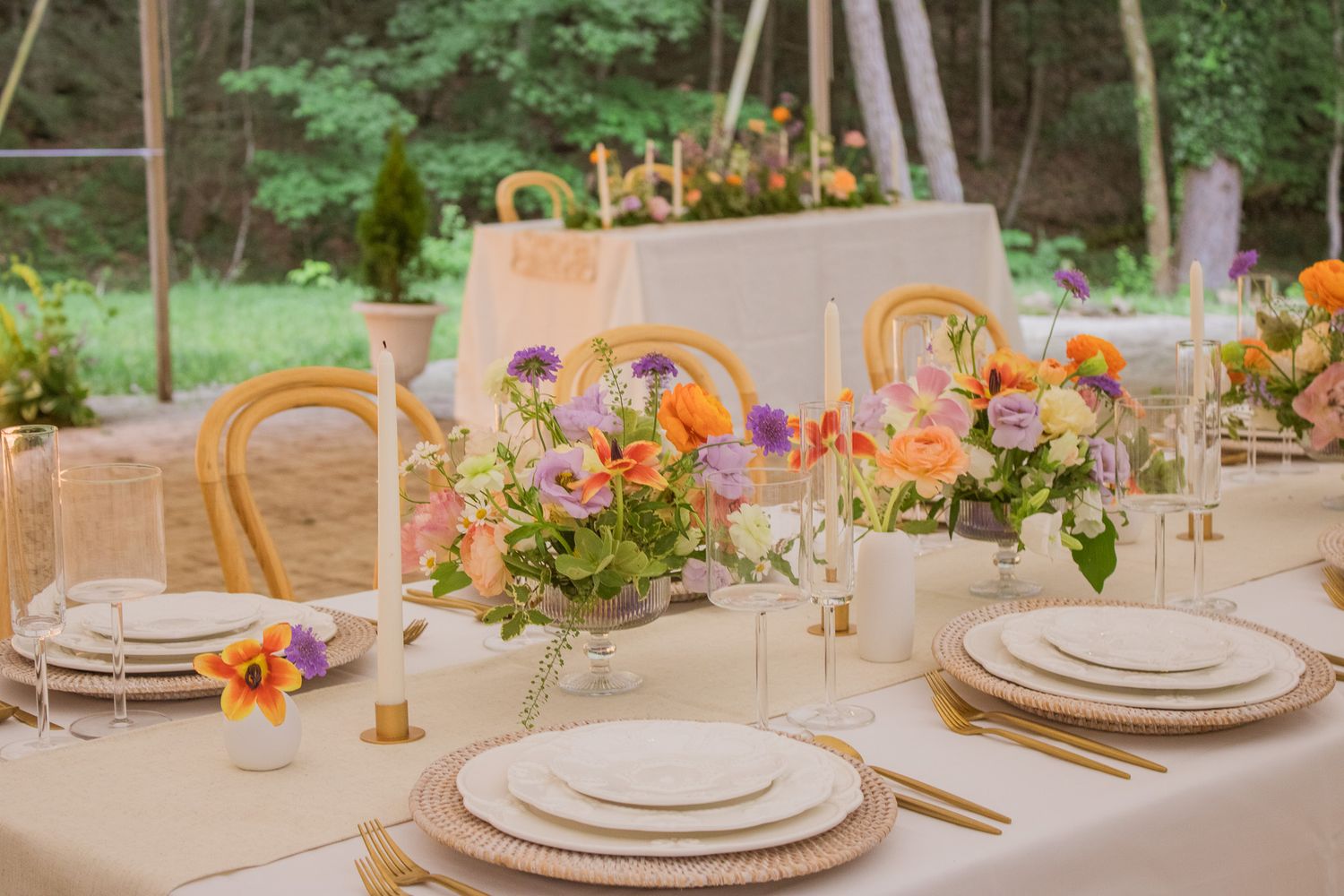 An elegant outdoor table setting features white plates, golden flatware, and colorful floral centerpieces in a garden.