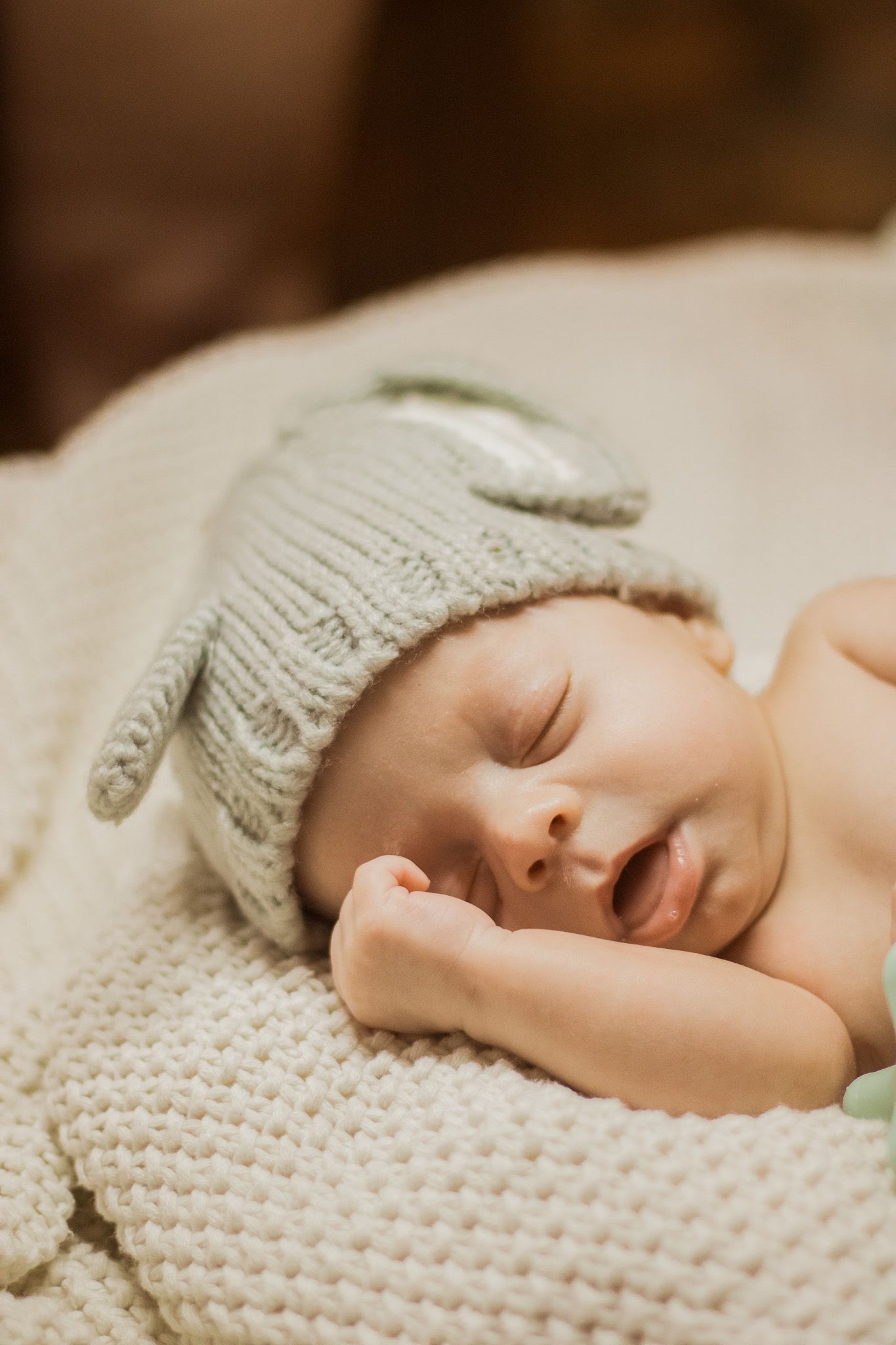 A sleeping newborn wearing a soft gray knit hat rests peacefully on a cream-colored textured blanket.