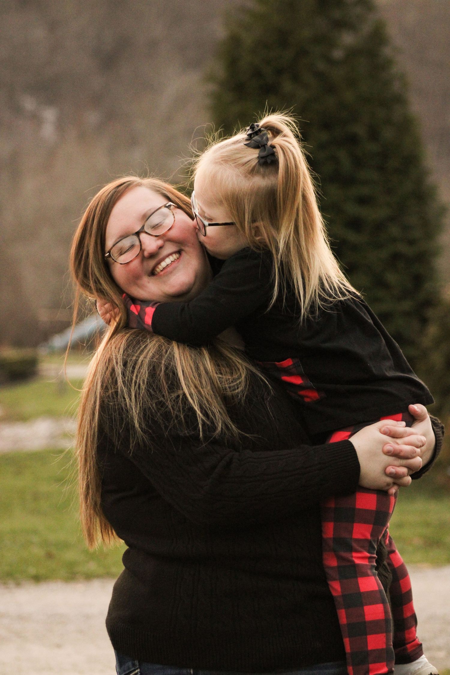 Two people share a joyful embrace outdoors with evergreen trees visible in the background.