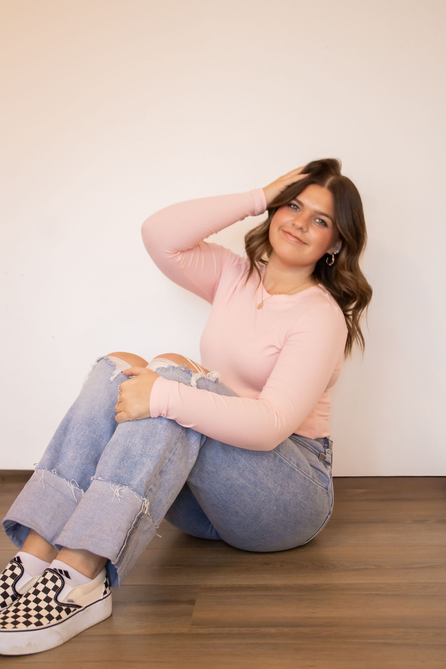 A person in a pink sweater and cuffed jeans sits casually on a hardwood floor wearing checkered slip-on shoes.
