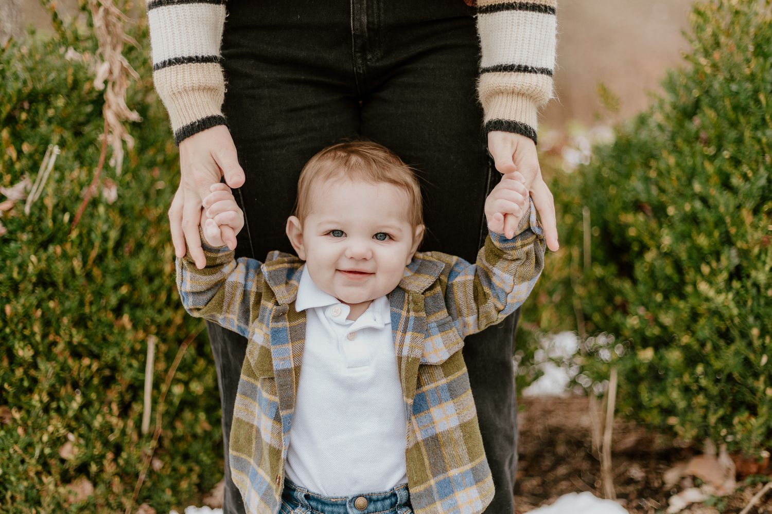 A toddler in plaid shirt and suspenders smiles while being held up outdoors against a natural green background.