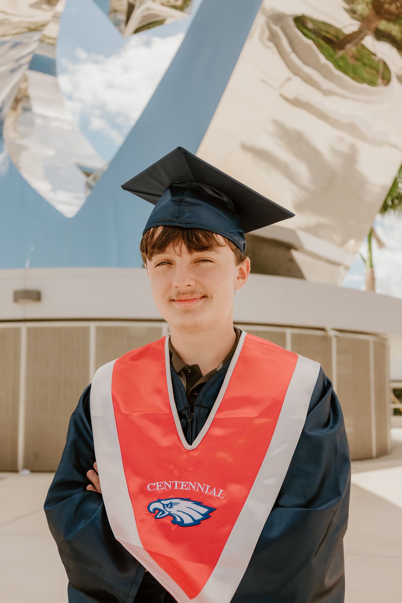 Graduate wearing black cap and gown with red Centennial sash stands outdoors on sunny day.
