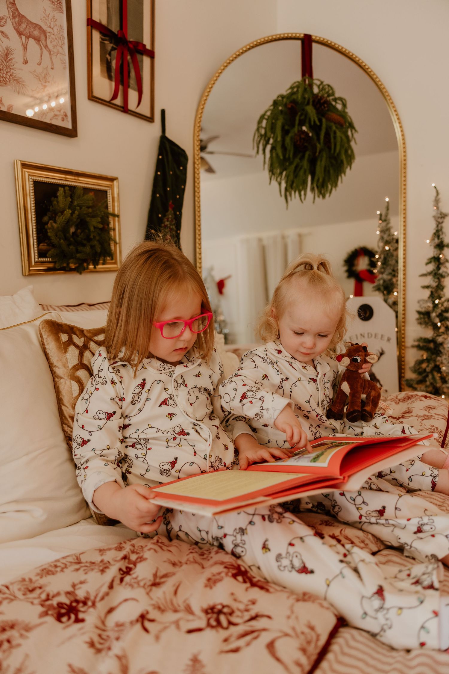 Two young children in matching Christmas pajamas read a storybook together on a festively decorated bed.