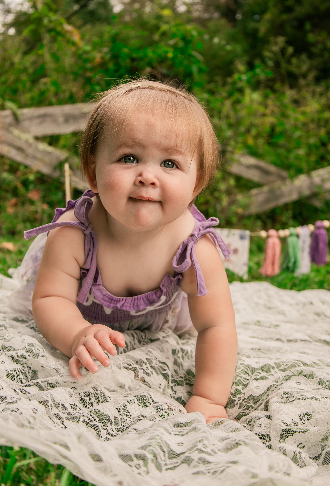 Baby in purple dress lying on white lace blanket with greenery in background.