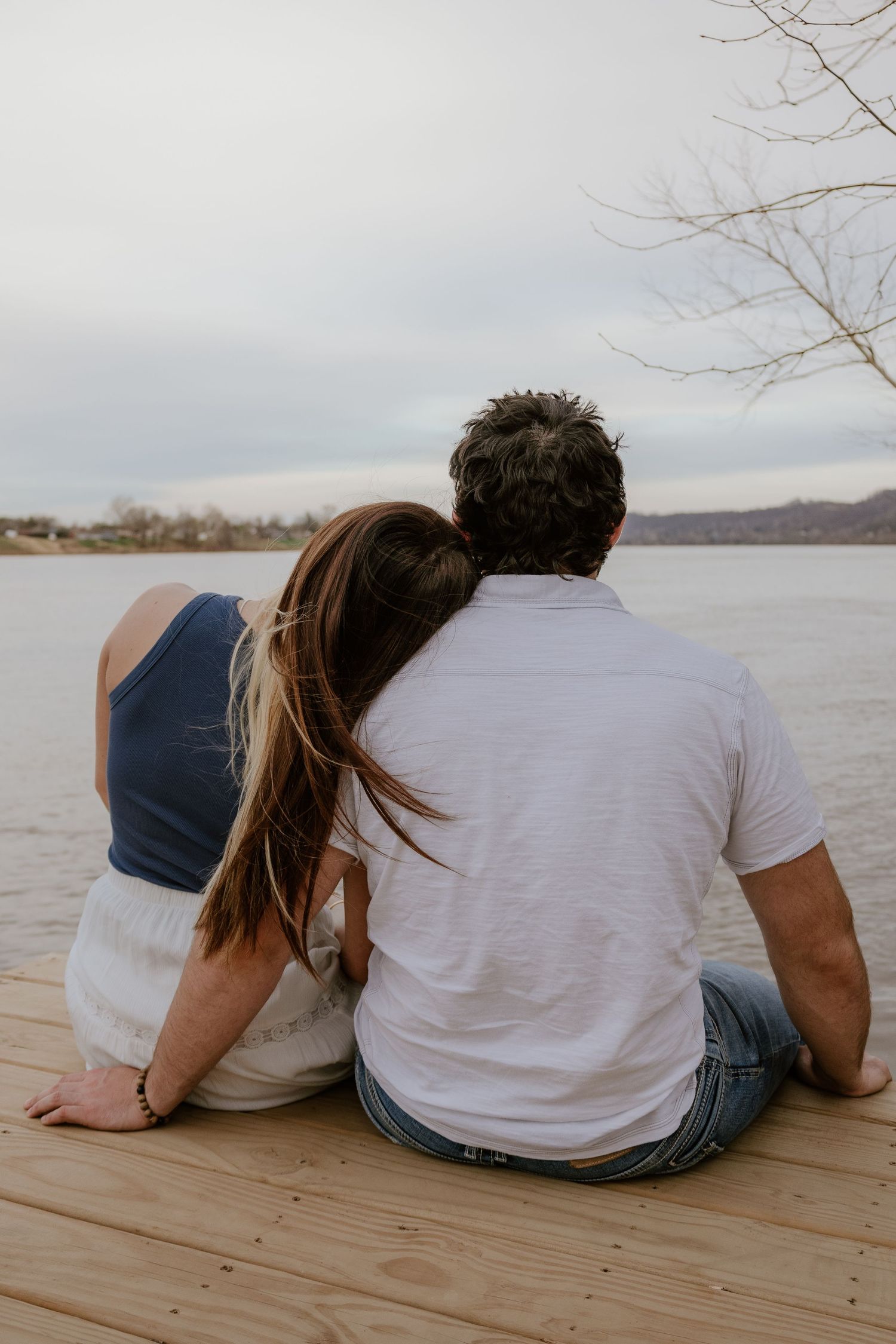 Two people sitting close together on a wooden dock by a lake on an overcast day wearing white shirts and jeans.