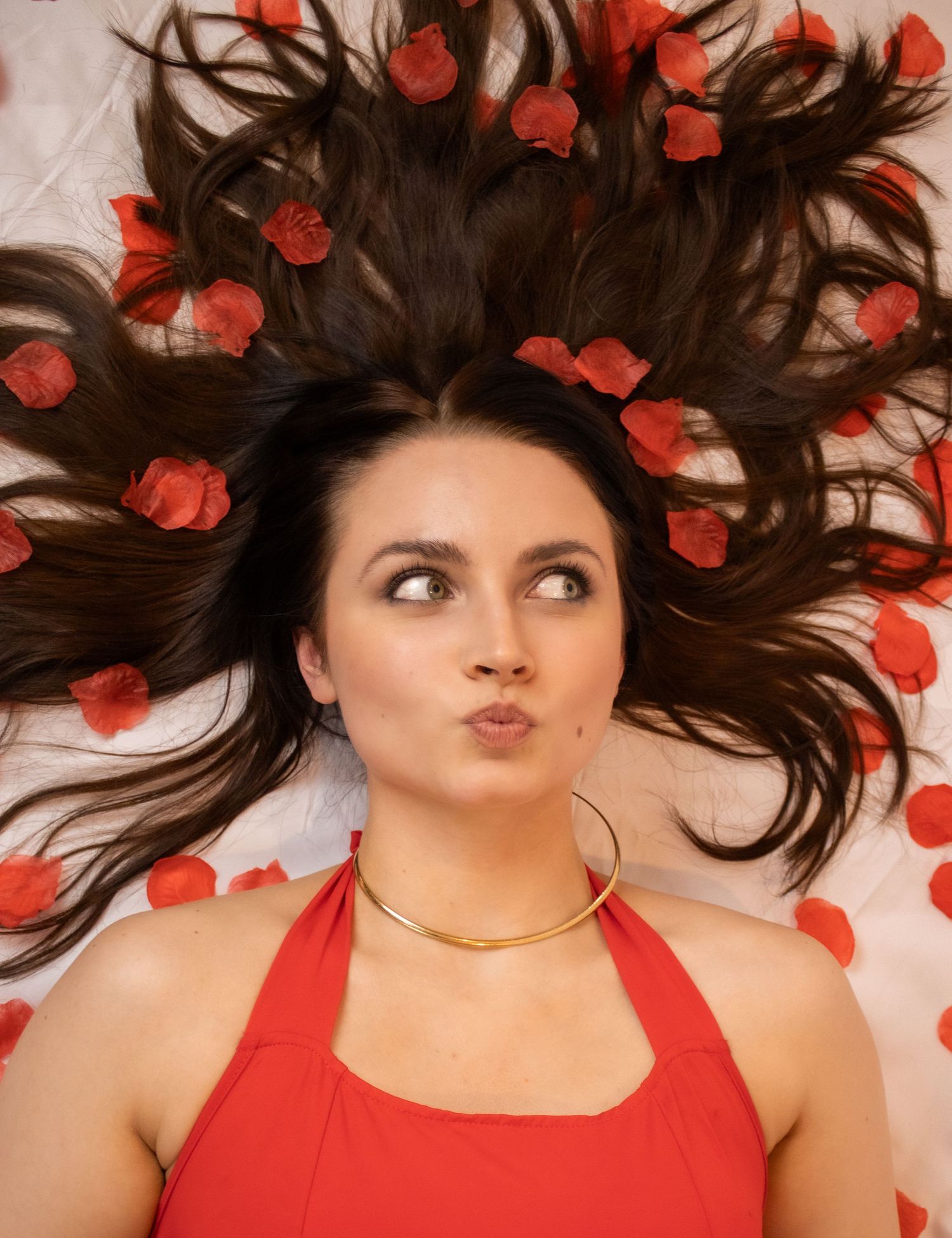 Woman in red tank top lying on white background with scattered red rose petals in flowing dark hair.