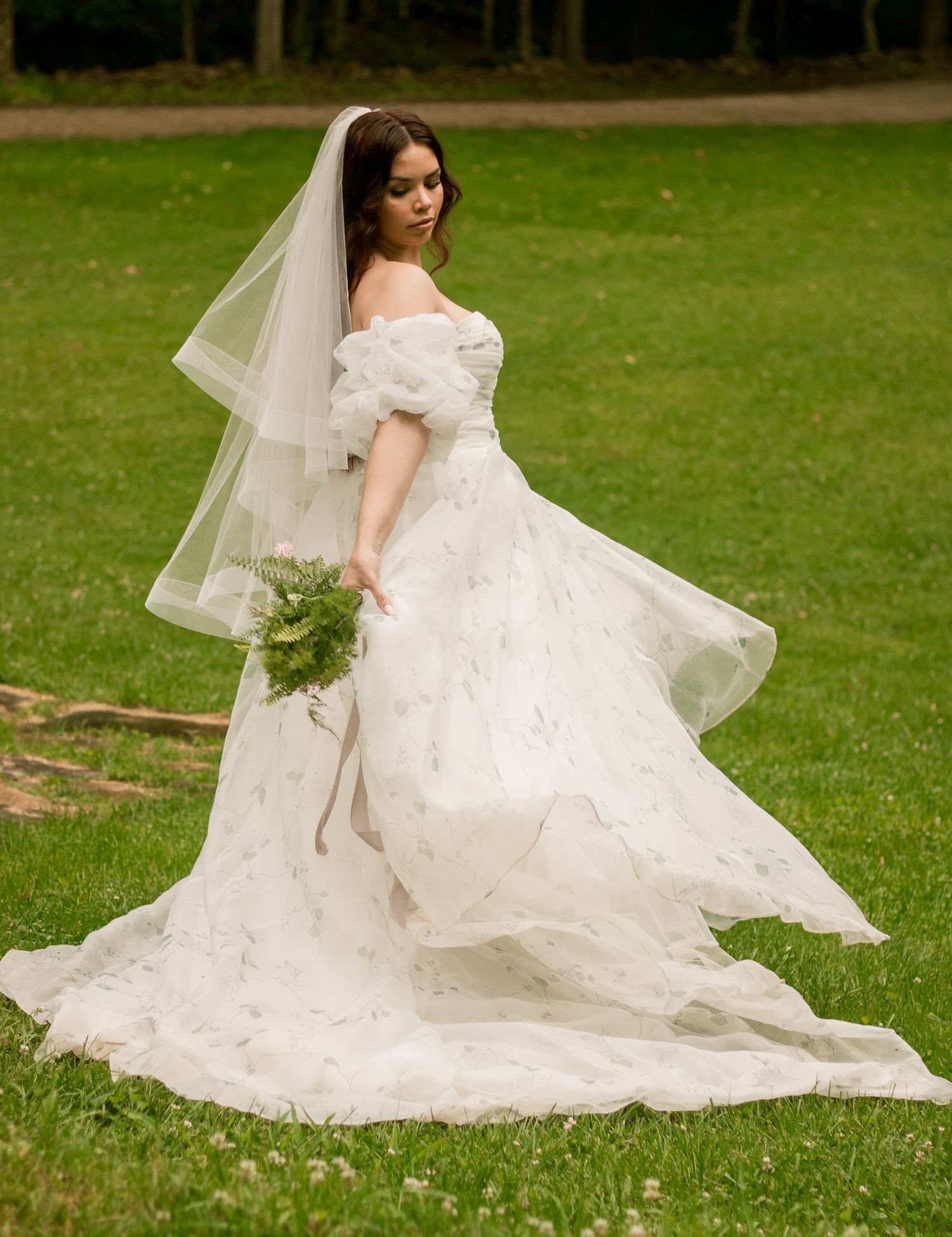 A bride in an off-shoulder lace wedding gown poses gracefully on a grassy lawn while holding a delicate bouquet.