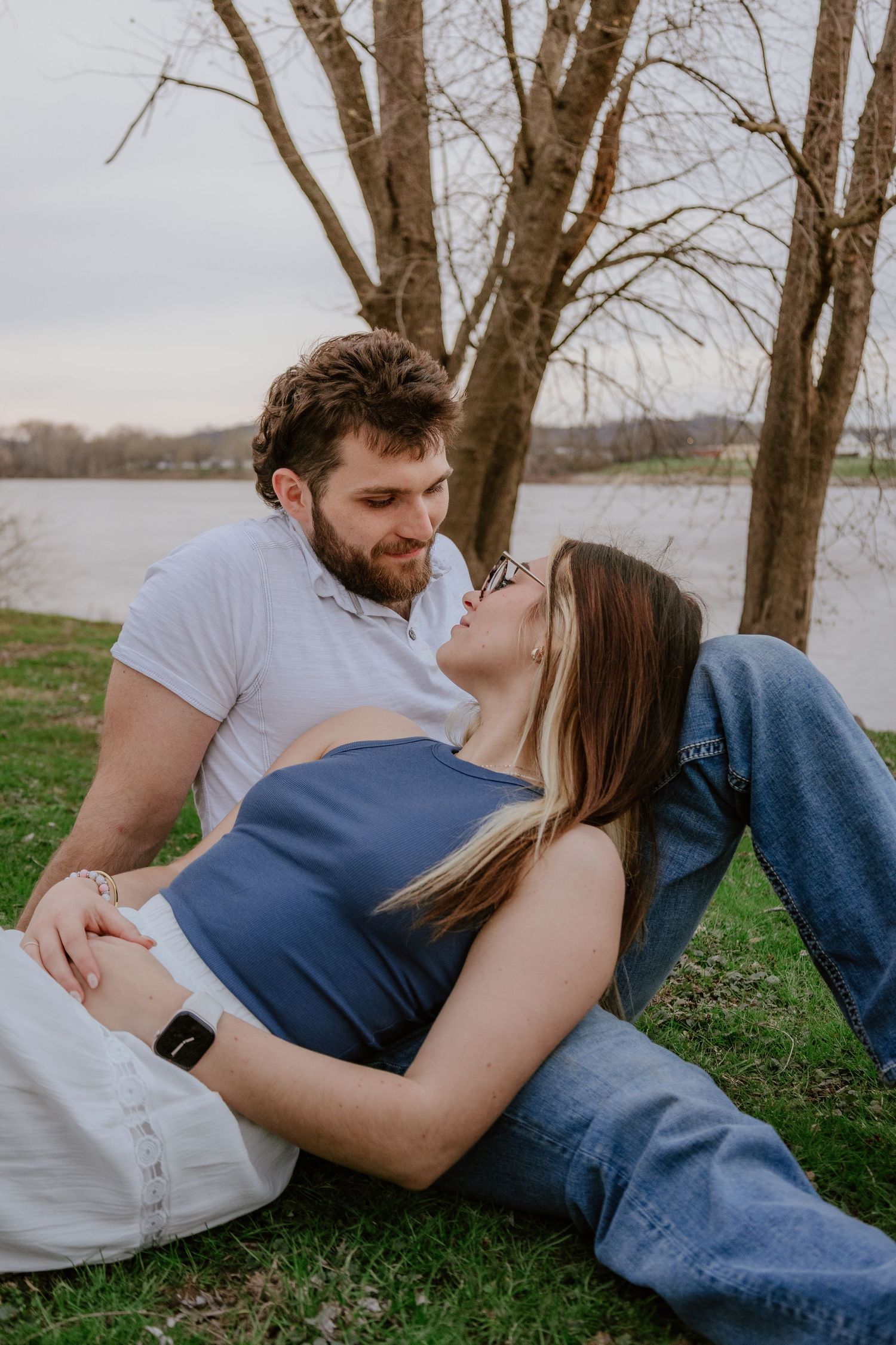 A couple in casual attire sharing an intimate moment while lying in grass near bare trees on an overcast day.