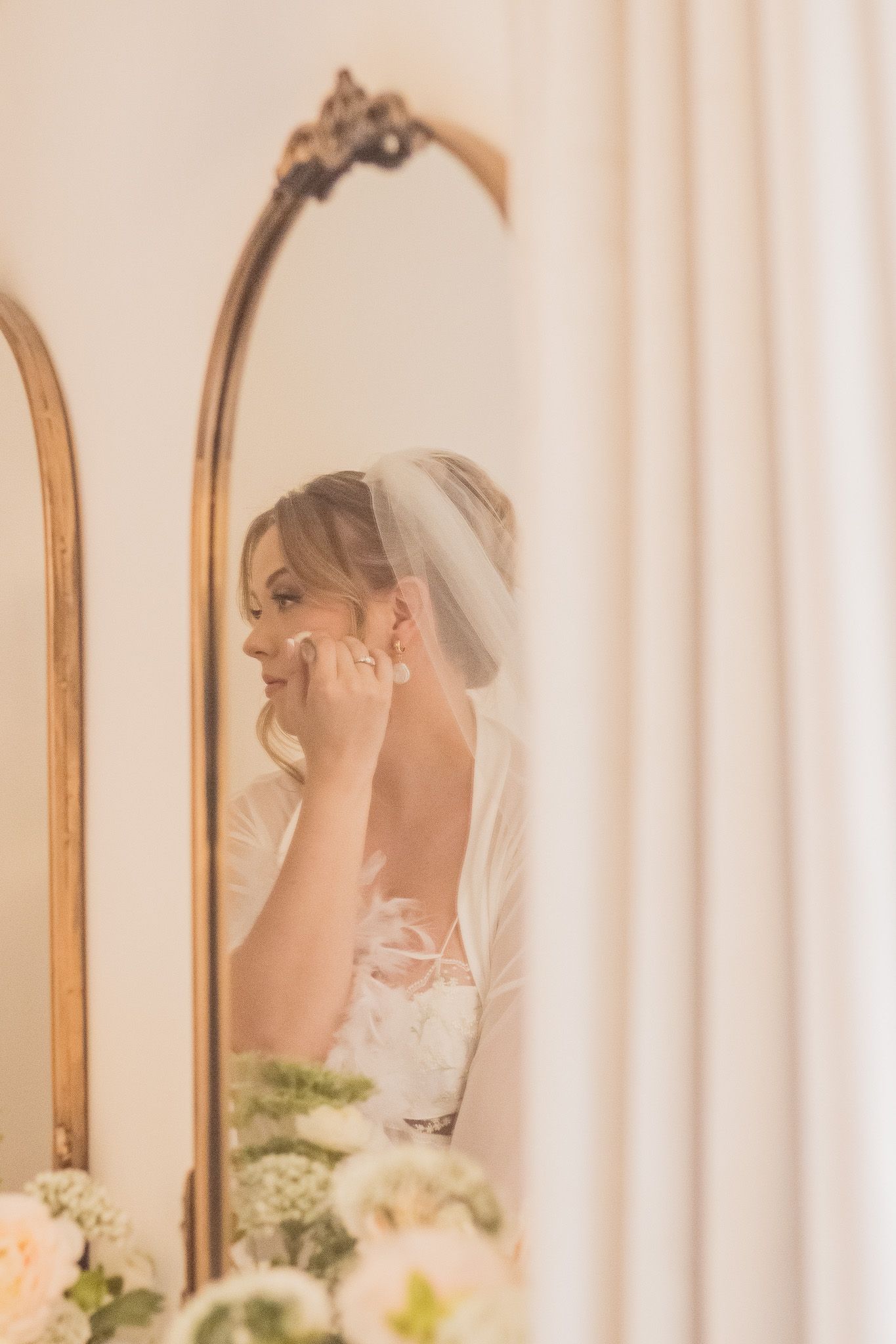 A bride in white lace veil is reflected multiple times in an elegant gold-framed mirror setup during wedding preparations.