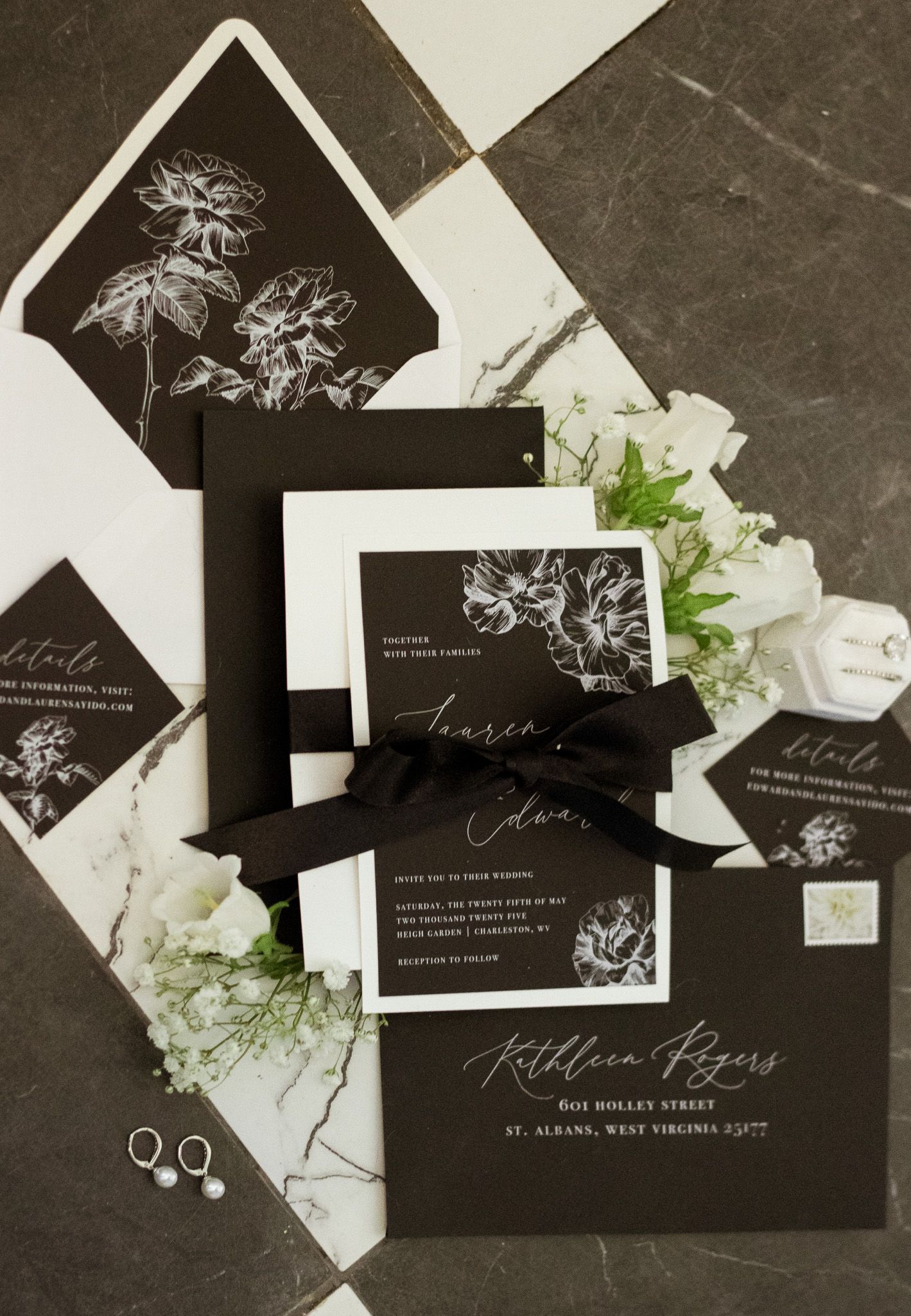 Black and white floral wedding invitation suite with white flowers and silver rings on dark surface.