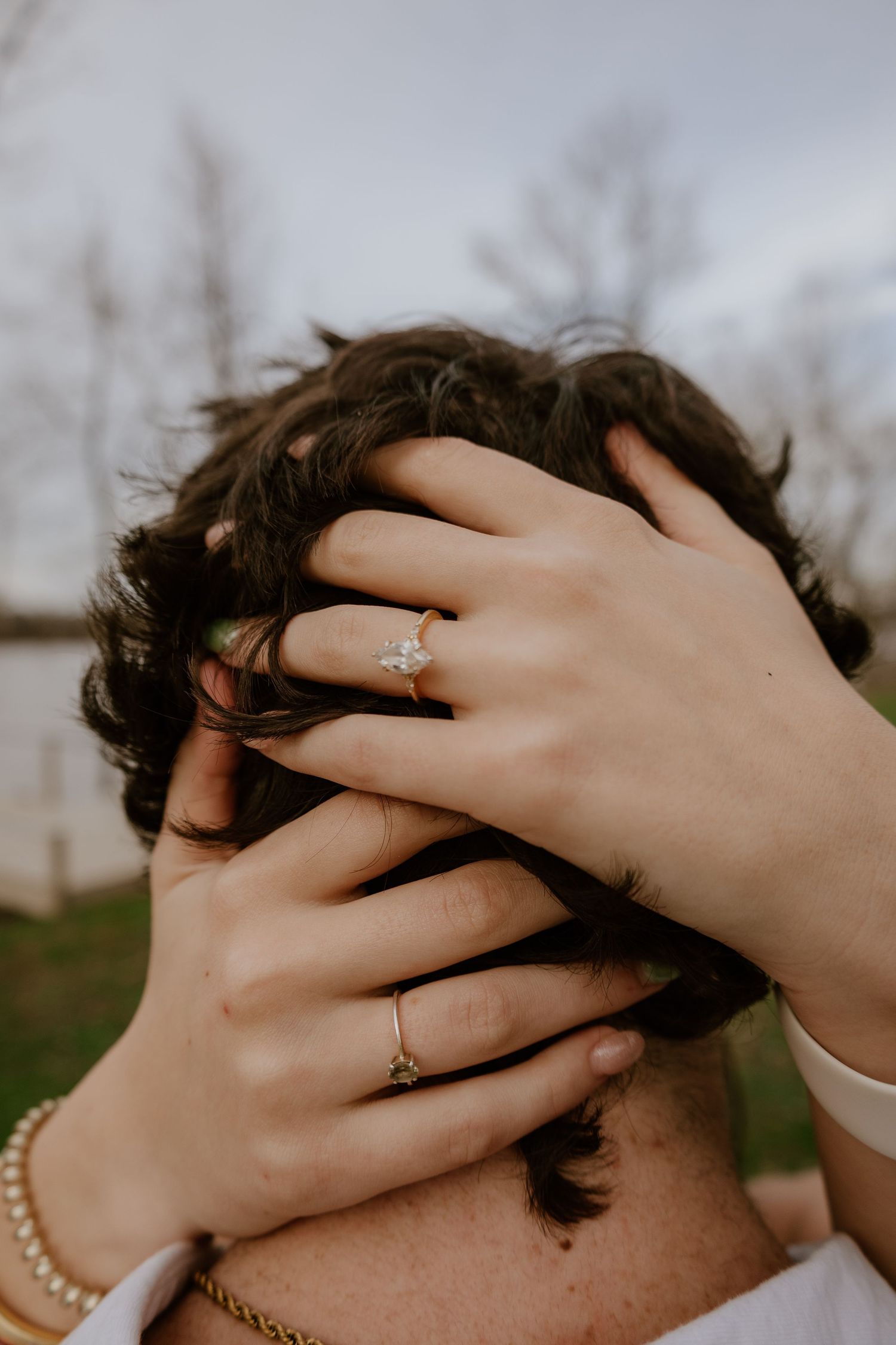 Hands with a delicate ring and pearl bracelet run through dark curly hair against a blurred outdoor background.