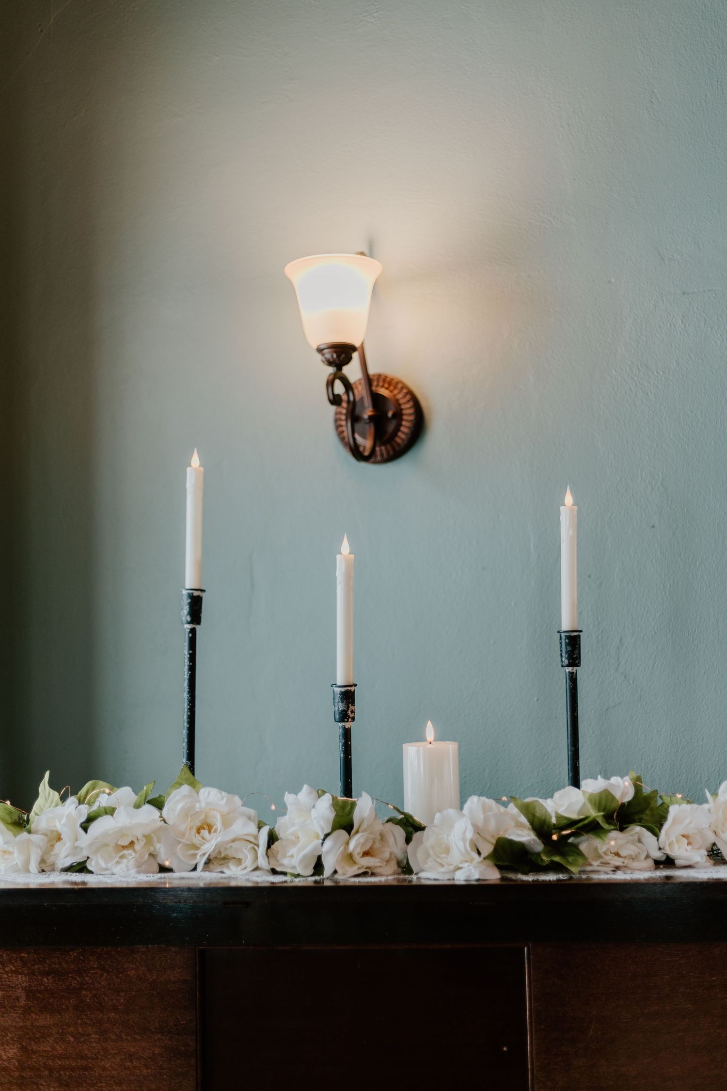Minimalist wedding decor features white candles and flowers arranged on dark wood against a soft blue wall.