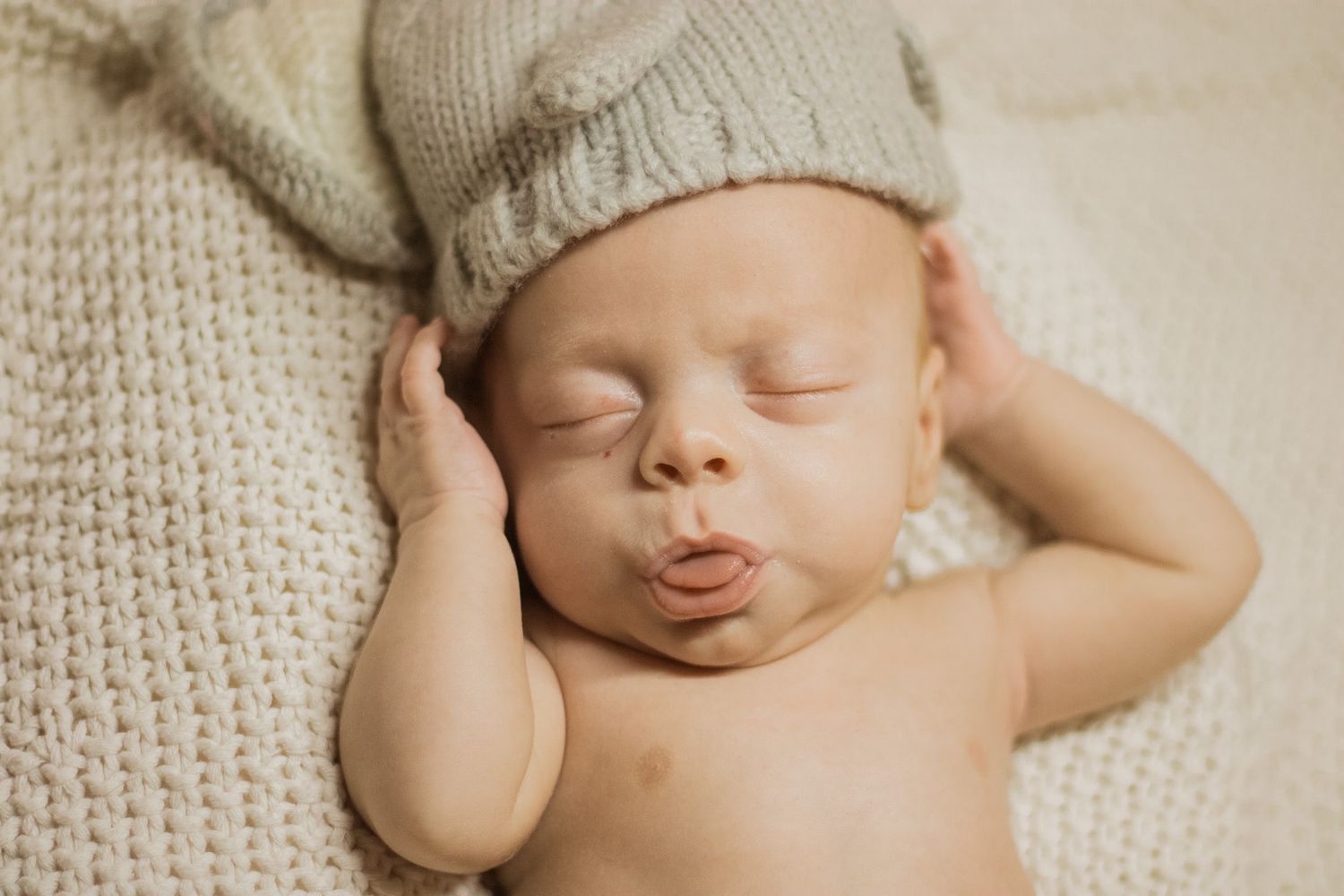 A sleeping newborn baby wearing a knit cap lies peacefully on soft white textured fabric.