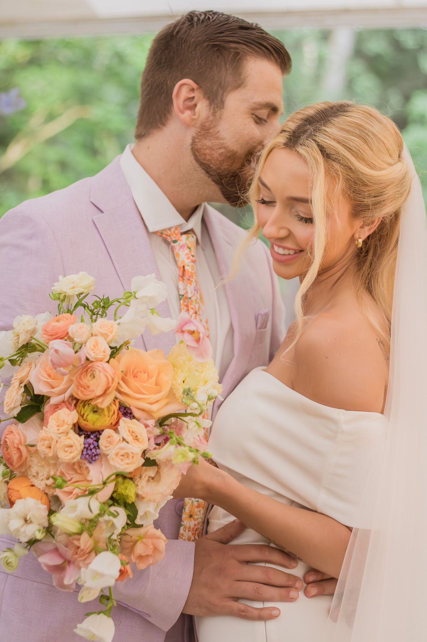 A romantic wedding couple embraces outdoors while holding a beautiful peach and blush floral bouquet in springtime.