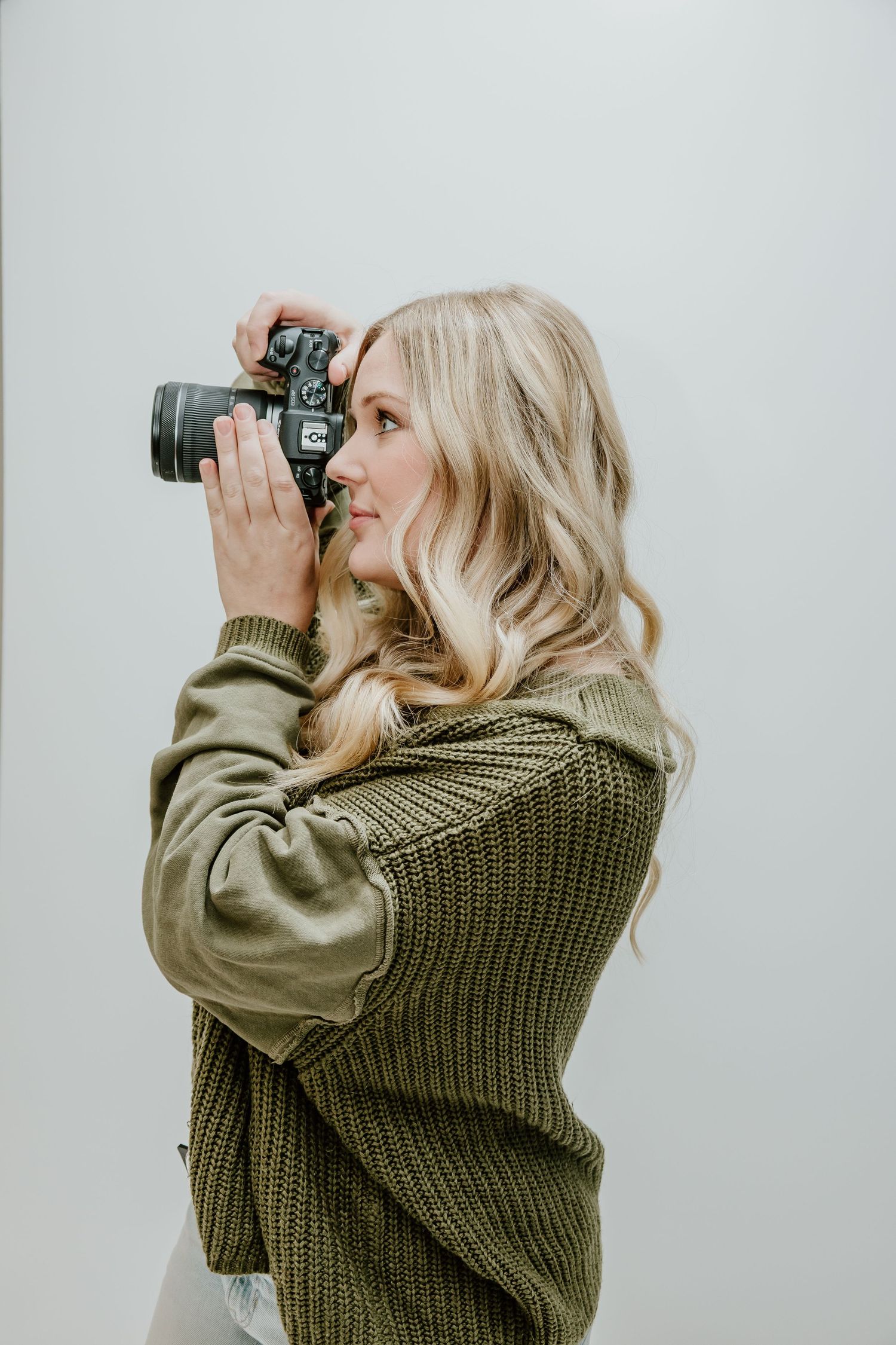 A photographer in an olive green sweater takes photos in profile view against a white background.