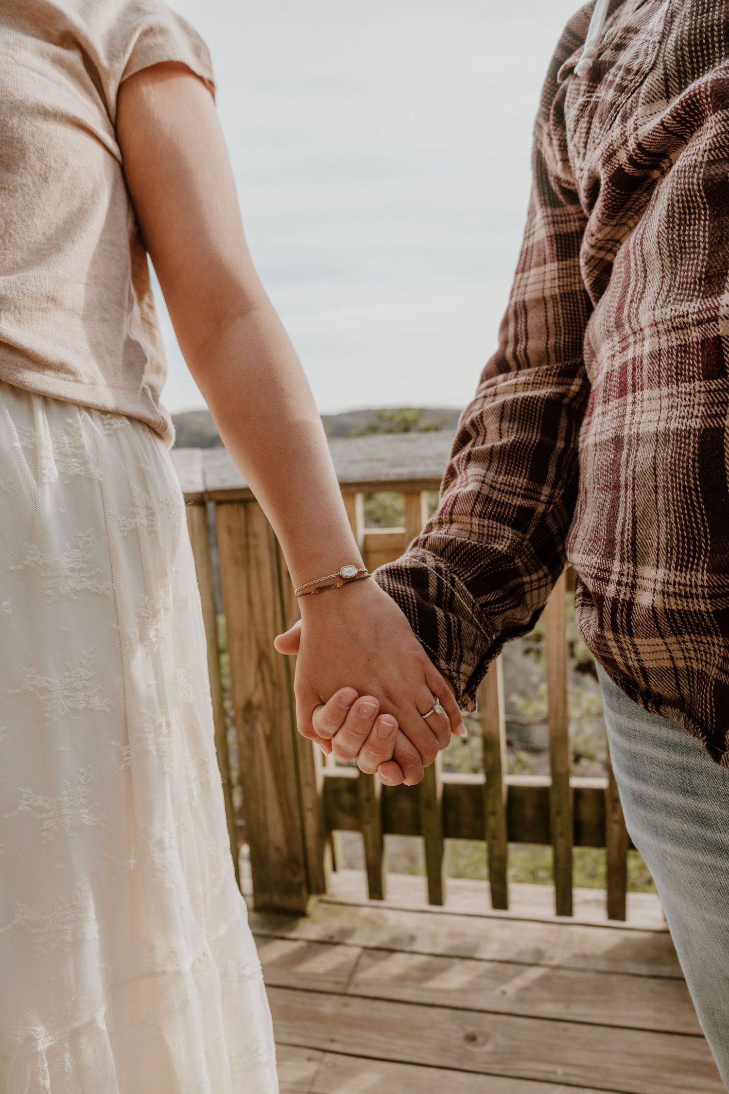 Close-up view of two people holding hands on a wooden deck overlooking a scenic landscape.