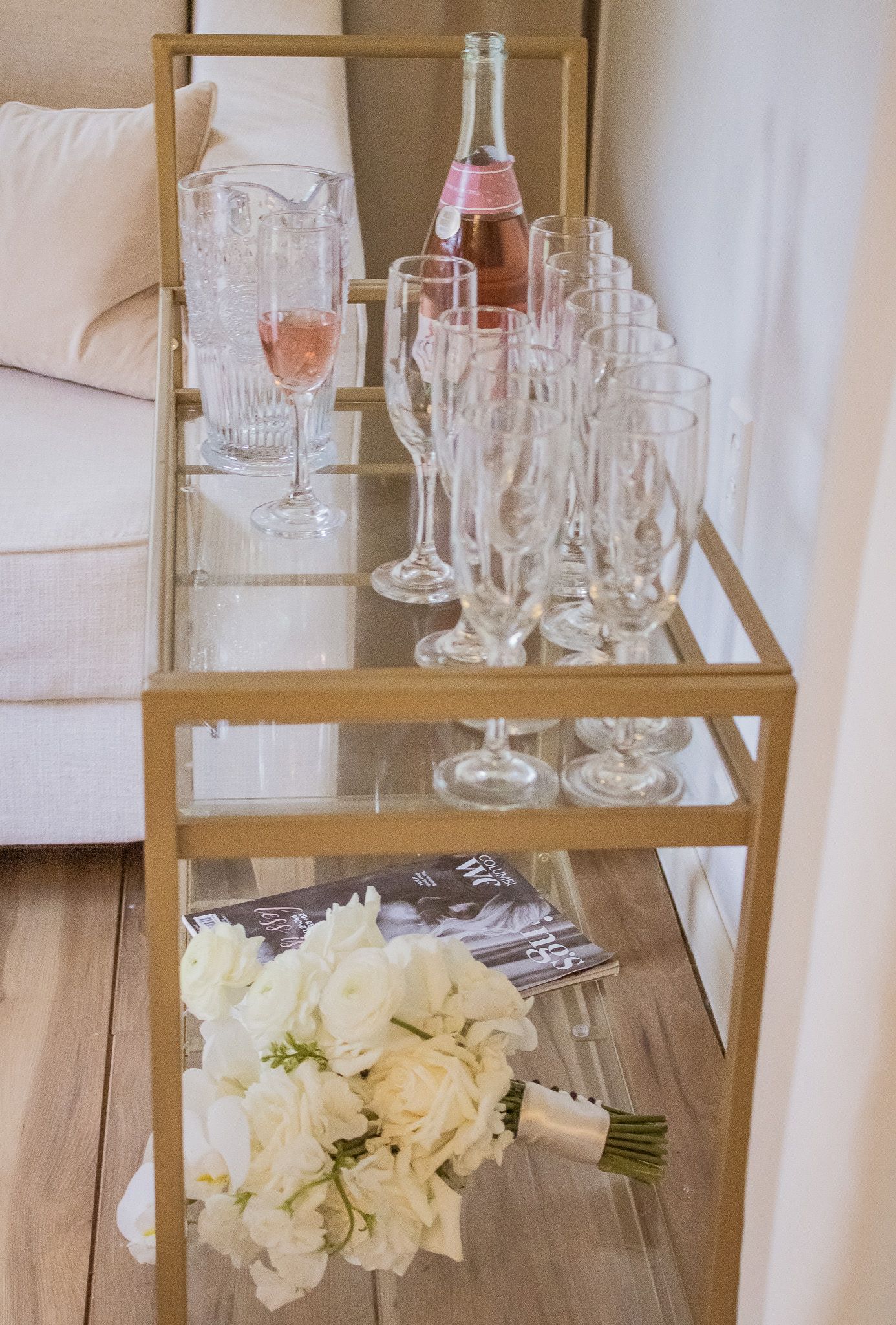 Gold bar cart with crystal champagne glasses, pink rosé wine, and white rose bouquet on wooden floor.