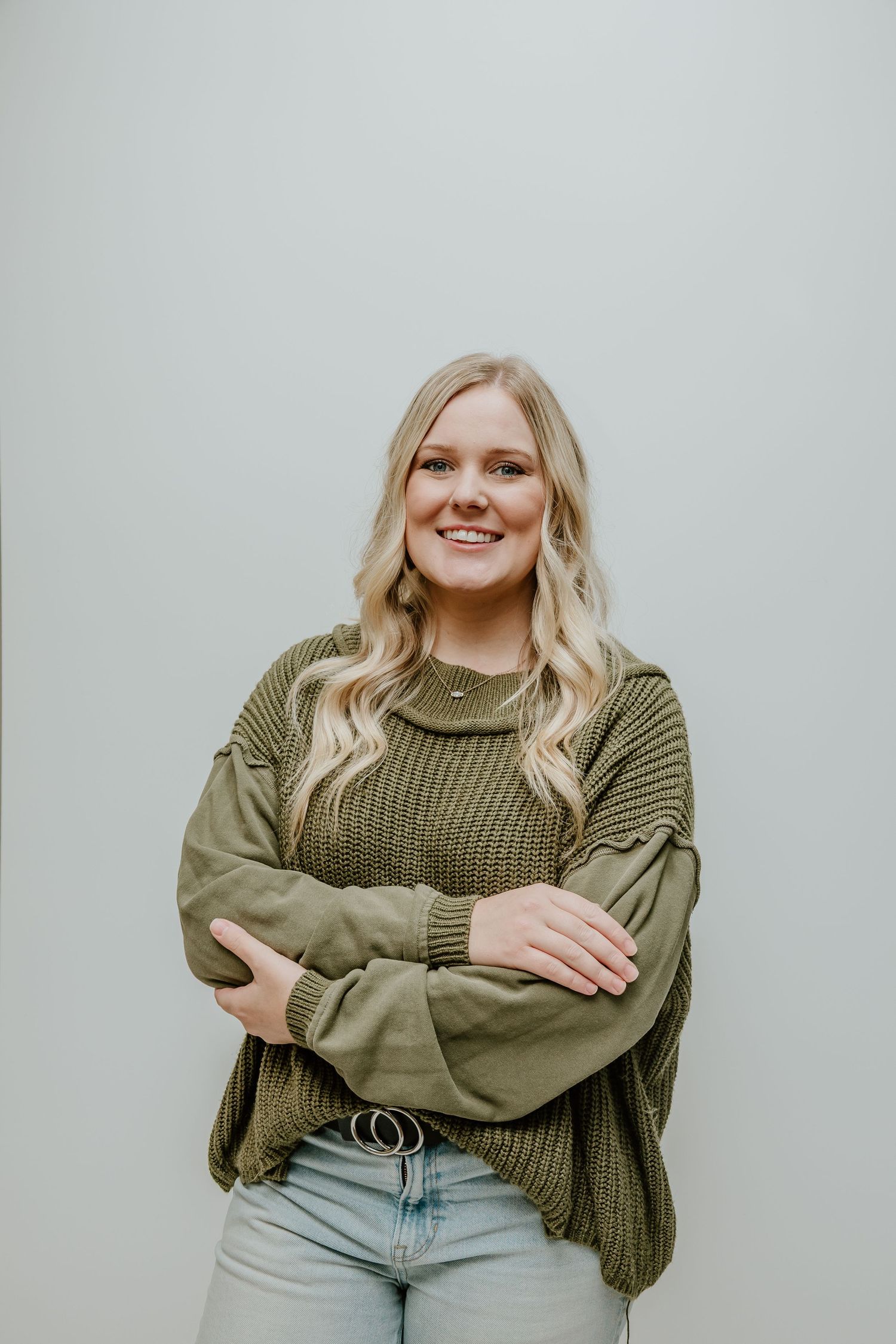 A person wearing an olive green knit sweater and light wash jeans poses against a gray background.