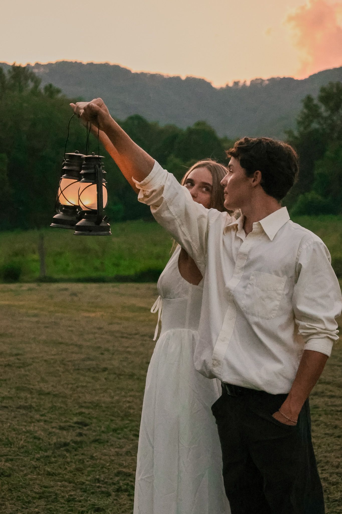 Two people in white shirts stand in a field at sunset holding lanterns up to the darkening sky.