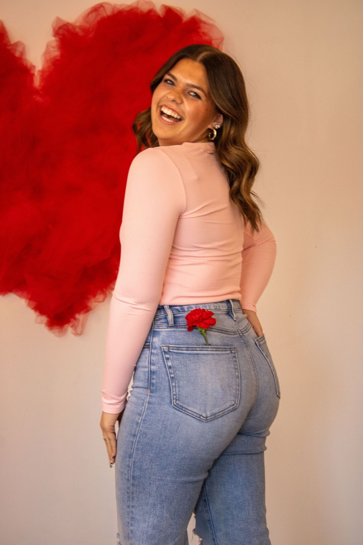 A joyful figure poses in a pink long-sleeve top and jeans against a red heart backdrop with a rose in back pocket.