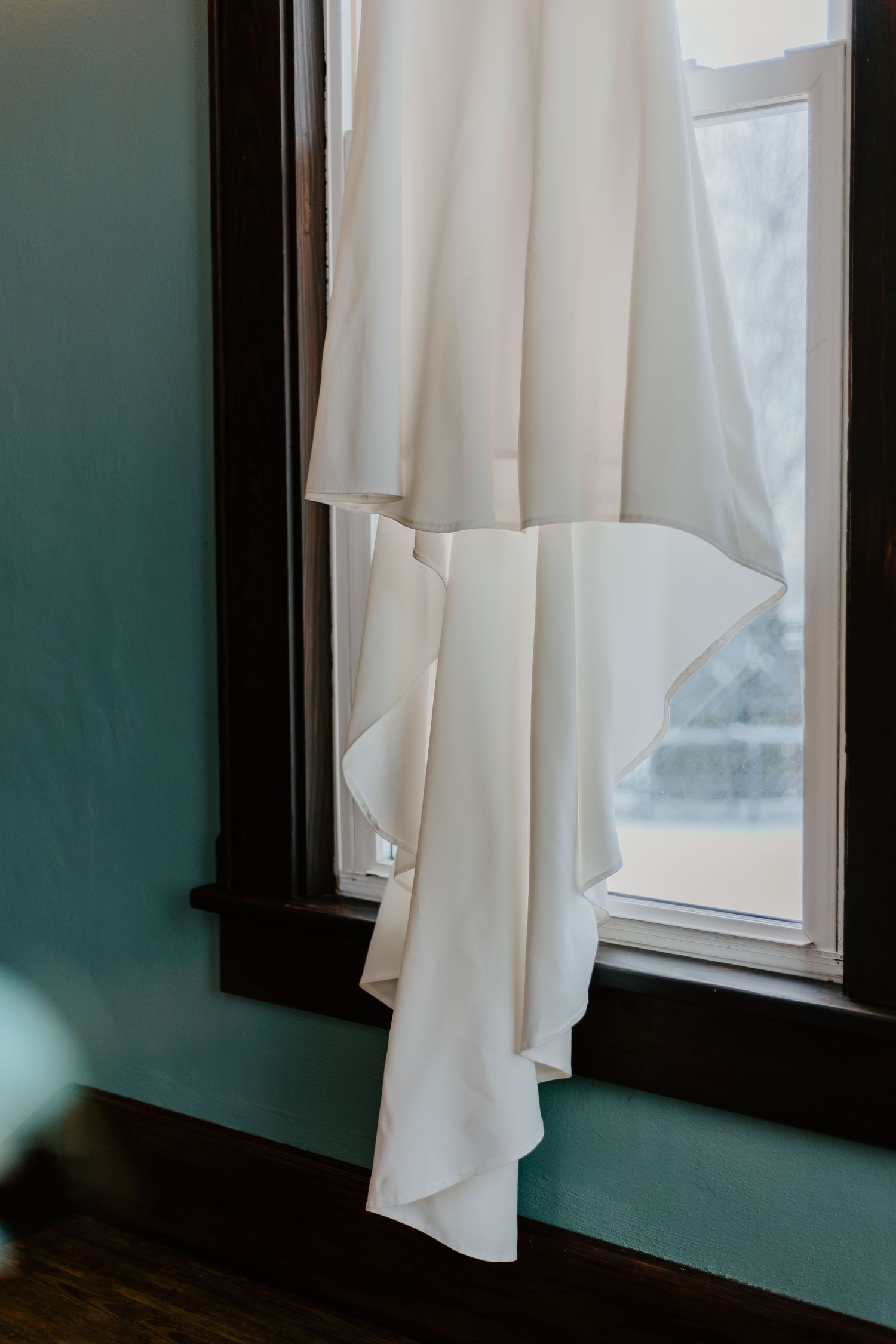 White wedding dress hang against turquoise walls near a window creating a soft, ethereal atmosphere.