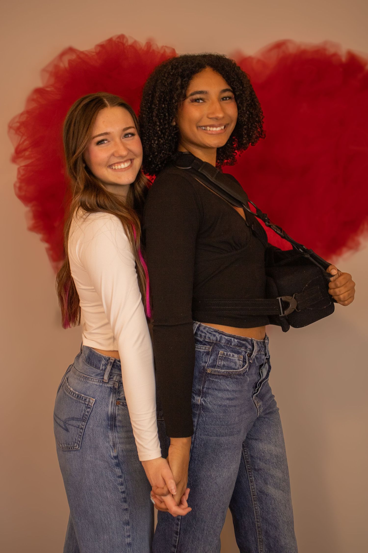 Two friends in jeans and contrasting tops pose playfully with red tulle props against a neutral backdrop.
