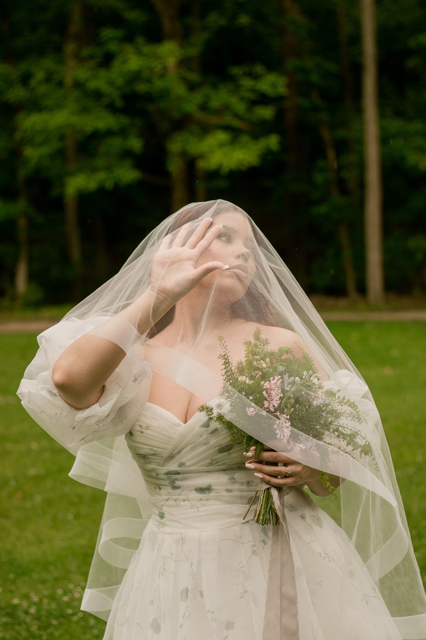 A dreamy bridal portrait showing a flowing veil and white dress against a backdrop of green trees.