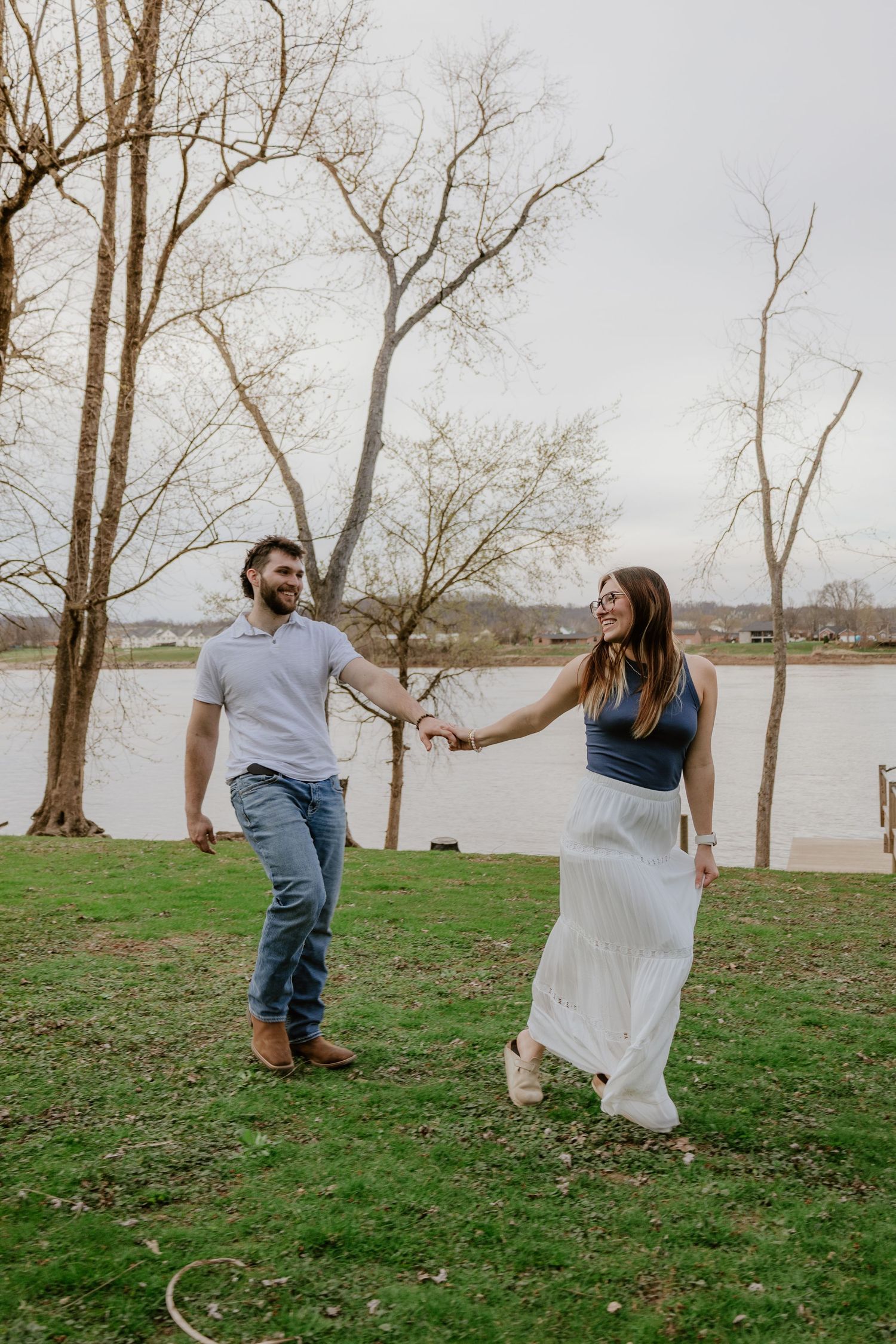 A couple dances playfully together on a grassy lakeside area with bare trees in the background during autumn.