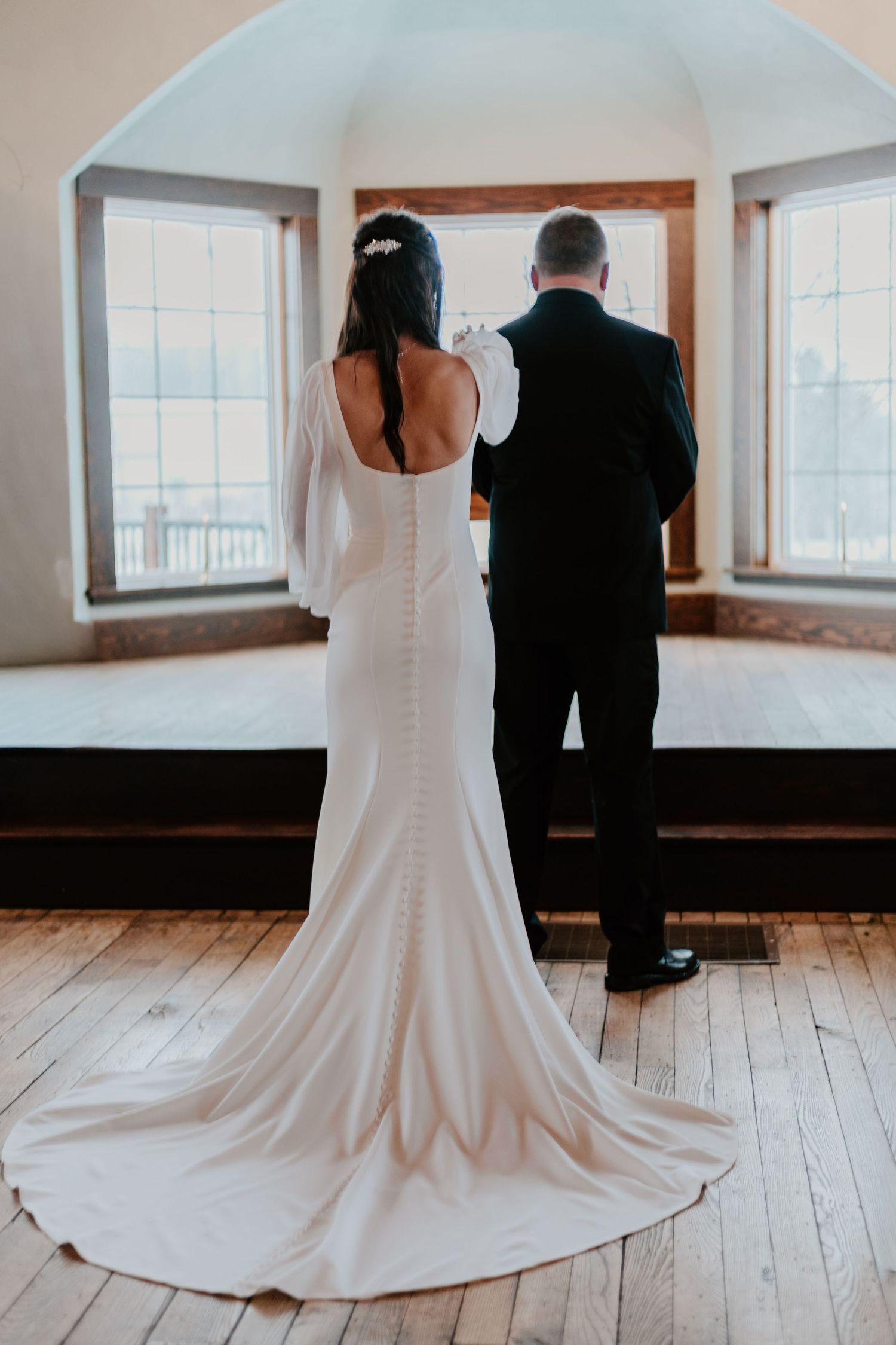 A bride in a white wedding dress and groom in black suit stand together on hardwood floors in a sunlit room.