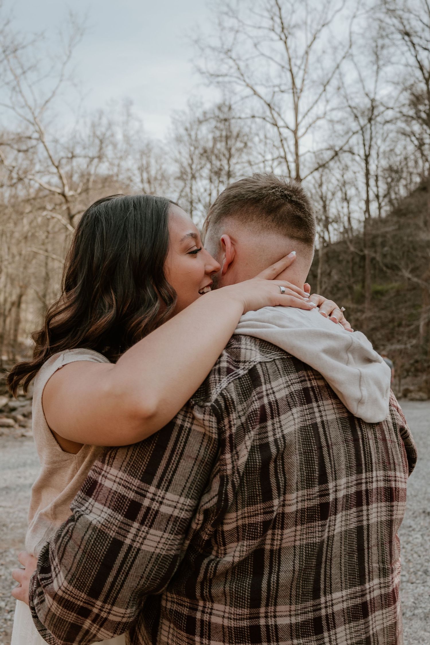 A romantic couple sharing a tender kiss outdoors in a wooded area wearing plaid and casual clothing.