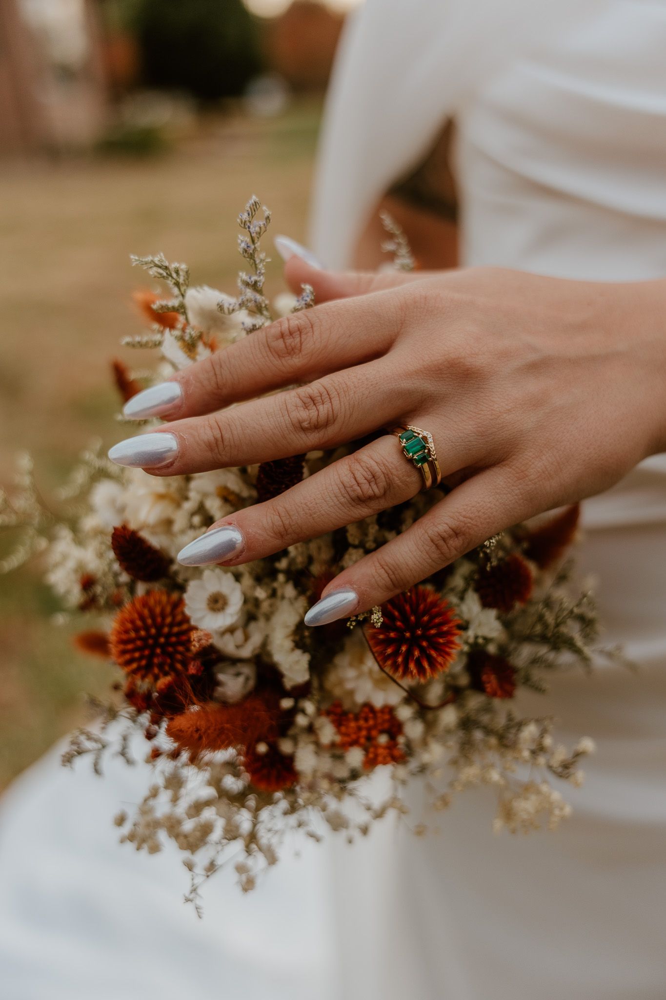 Close-up of hands with white nails and turquoise ring holding rustic autumn wedding bouquet with orange and dried flowers.