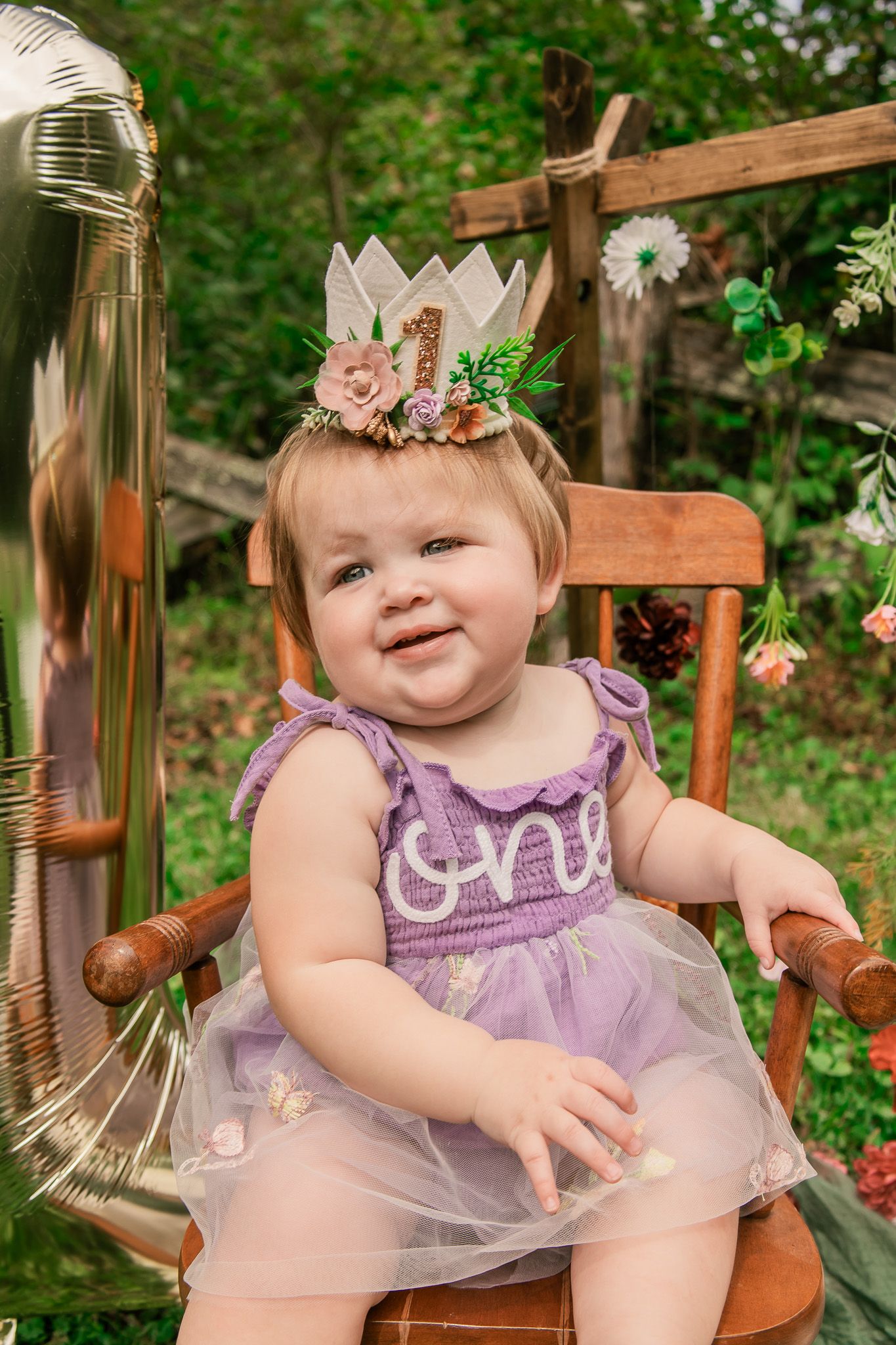 Baby wearing purple tulle dress and floral crown sits in wooden chair in garden setting.