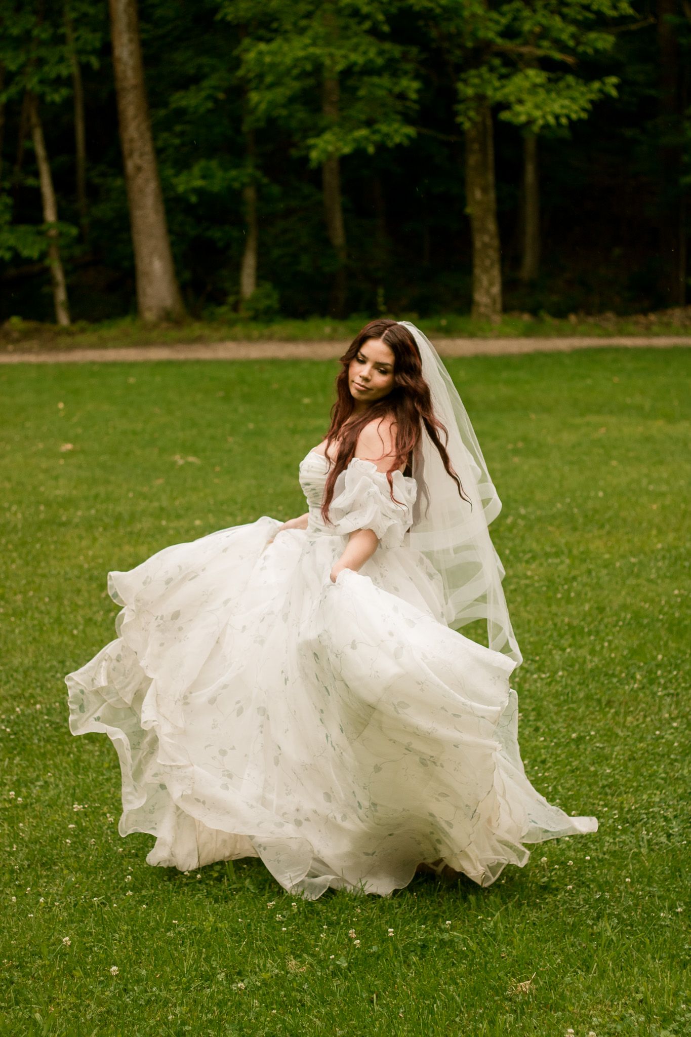 A bride in a flowing white wedding dress sits gracefully on lush green grass in a wooded setting.