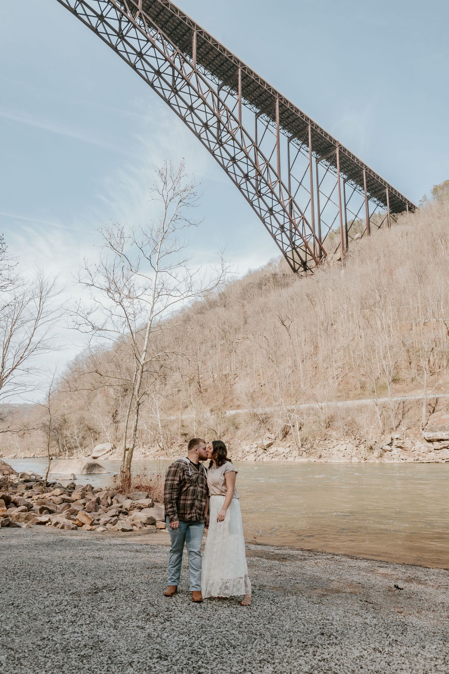 Couple standing beneath a towering steel bridge structure against a winter landscape with bare trees.