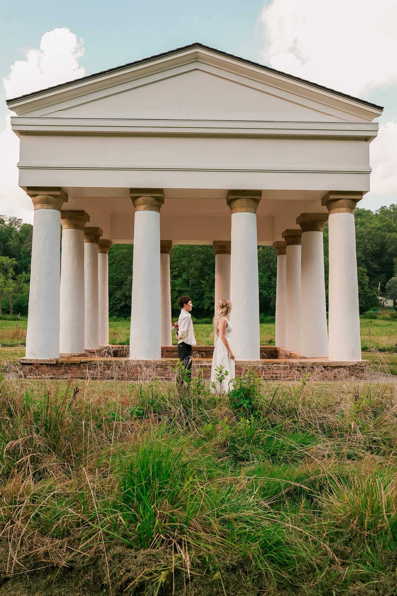 A classical columned pavilion with white pillars stands in a grassy field while two people stand beneath its architecture.