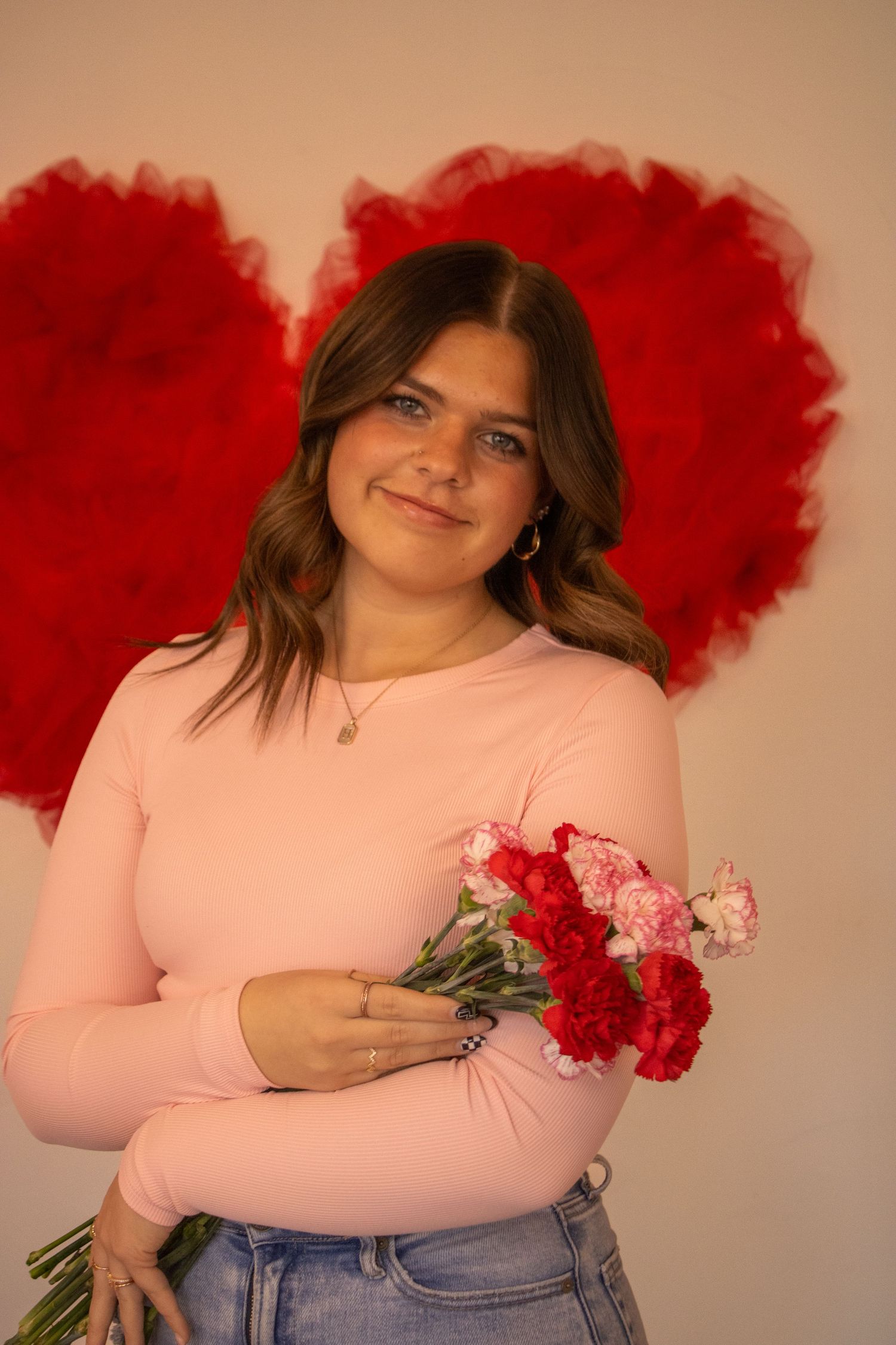 A person in a pink sweater holds red and pink carnations while posing in front of a painted red heart backdrop.